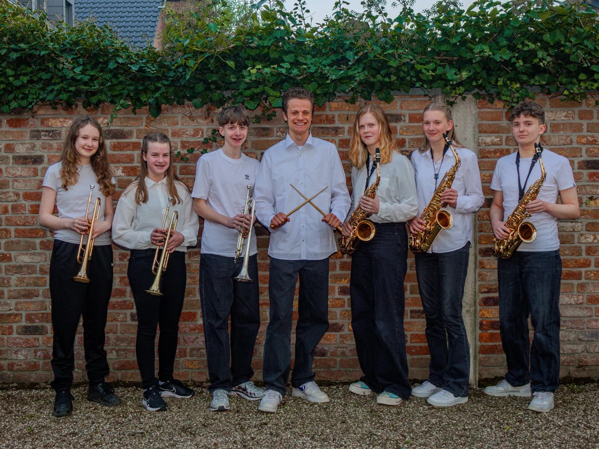 Gruppe von sieben Personen mit Musikinstrumenten vor einer Backsteinmauer. Drei Trompeten und drei Saxophone sind zu sehen. Eine Person in der Mitte hält Trommelstöcke.
