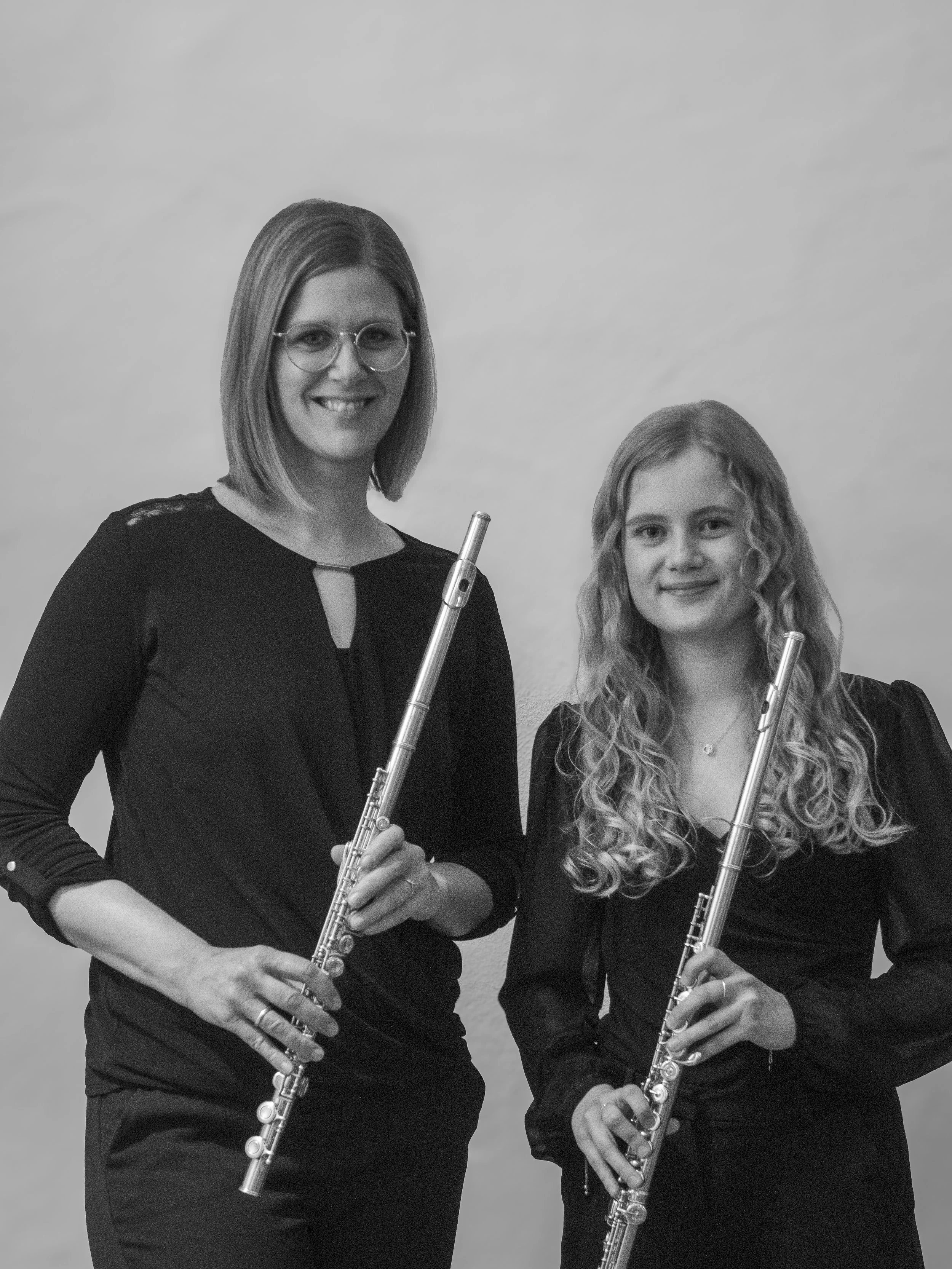 Zwei Frauen, die Querflöten halten, lächeln in die Kamera. Beide tragen schwarze Kleidung und stehen vor einem neutralen Hintergrund.