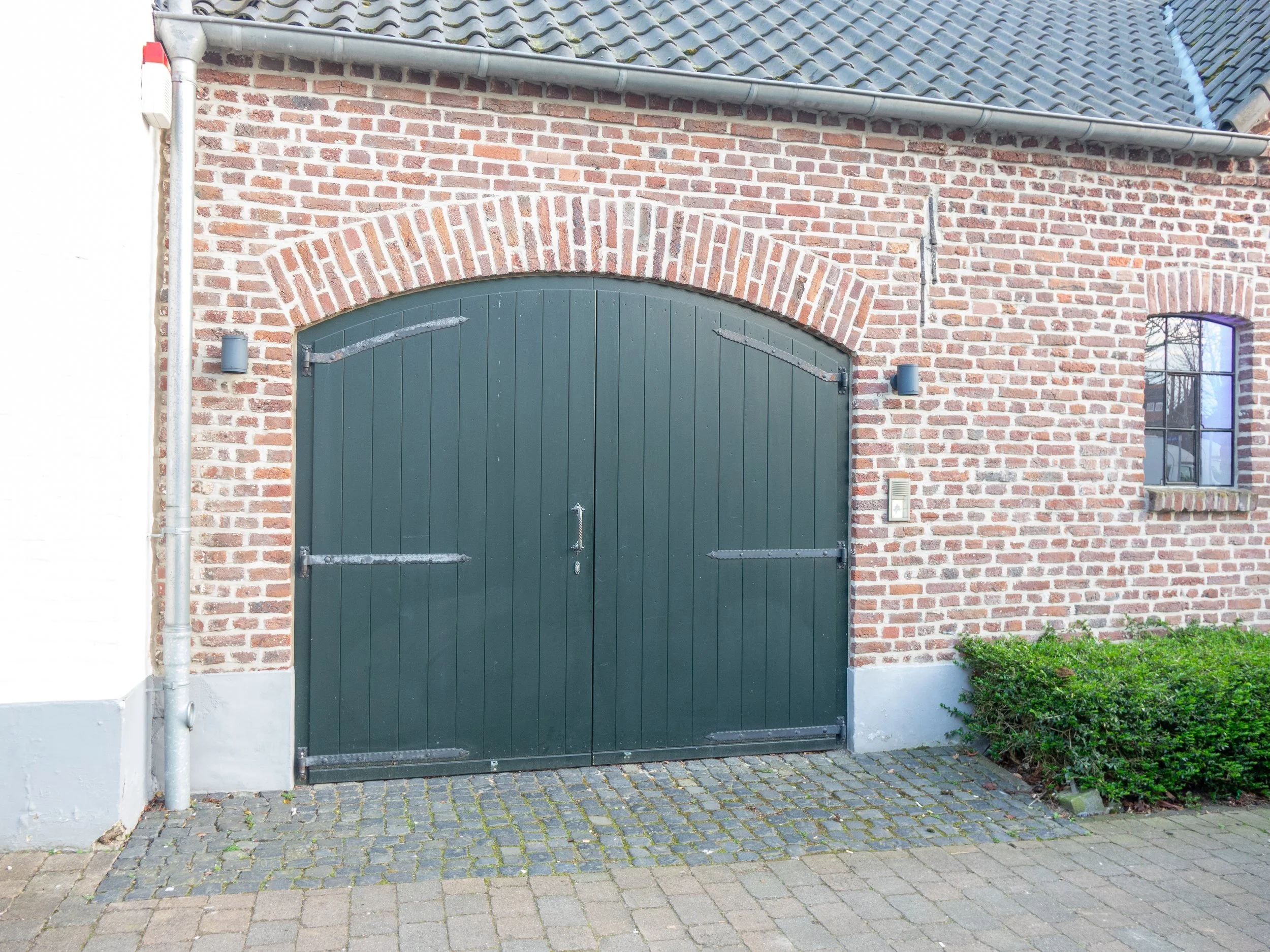 Große grüne Holztor in einer Backsteinfassade mit einem kleinen Fenster rechts und einem gepflasterten Gehweg davor.