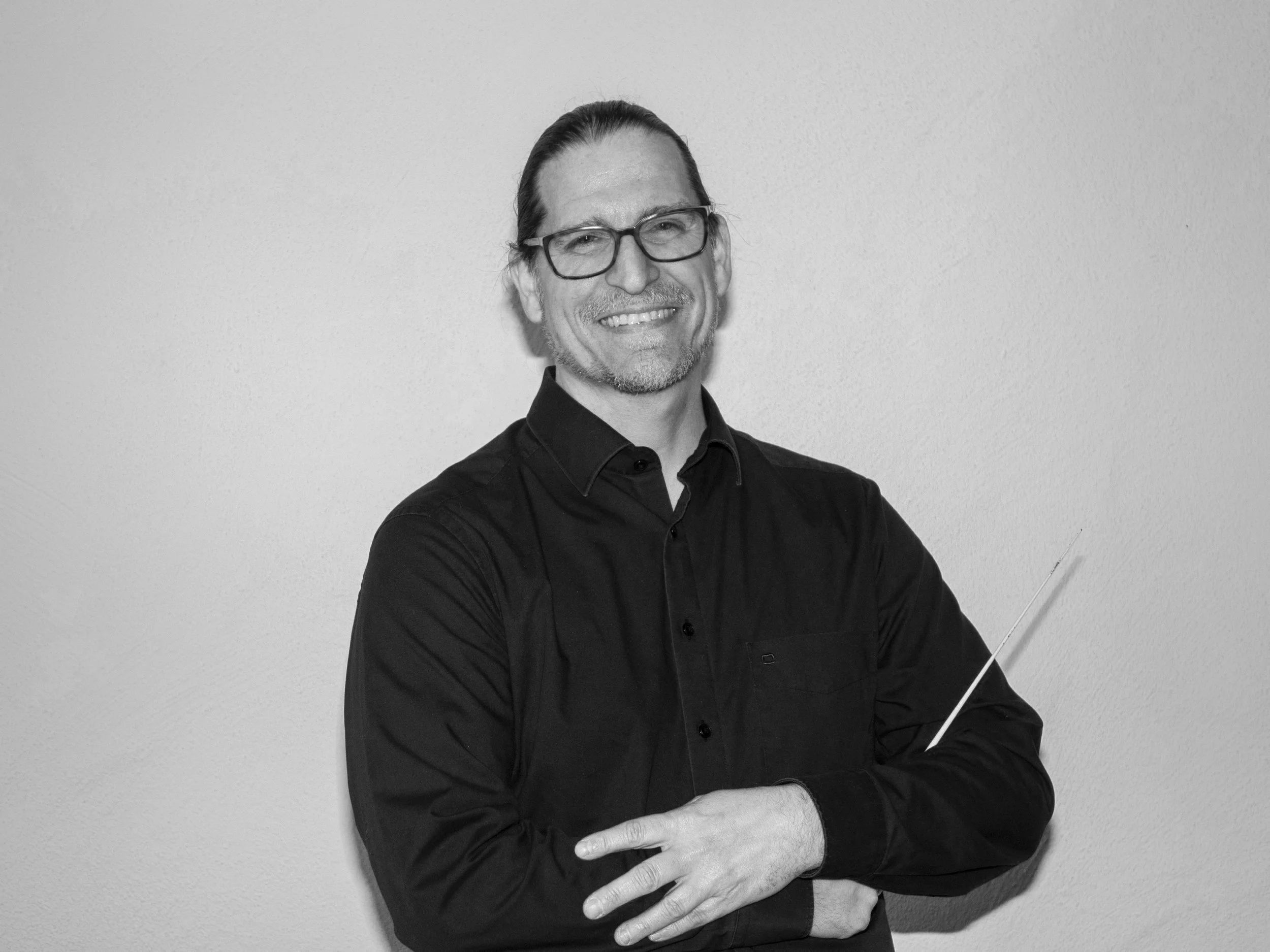 Ein lächelnder Mann in schwarzem Hemd mit Brille und zusammengebundenem Haar, hält einen Dirigentenstab.