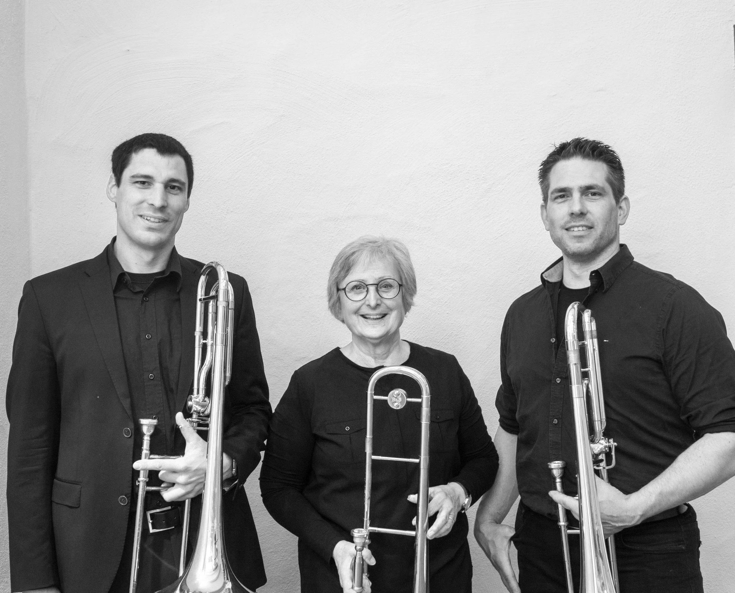 Drei Personen stehen nebeneinander und halten Posaunen, lächelnd in Schwarz-Weiß fotografiert.