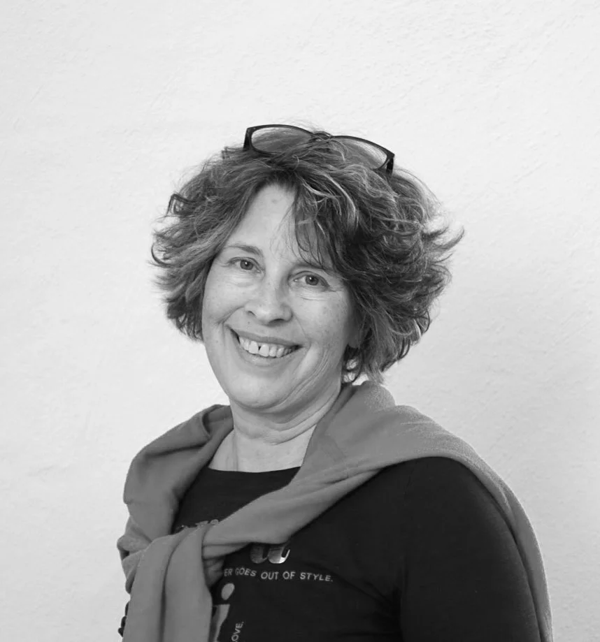 Schwarzweißfoto einer lächelnden Person mit lockigem Haar. Sie trägt ein dunkles Oberteil und hat eine Brille auf dem Kopf.