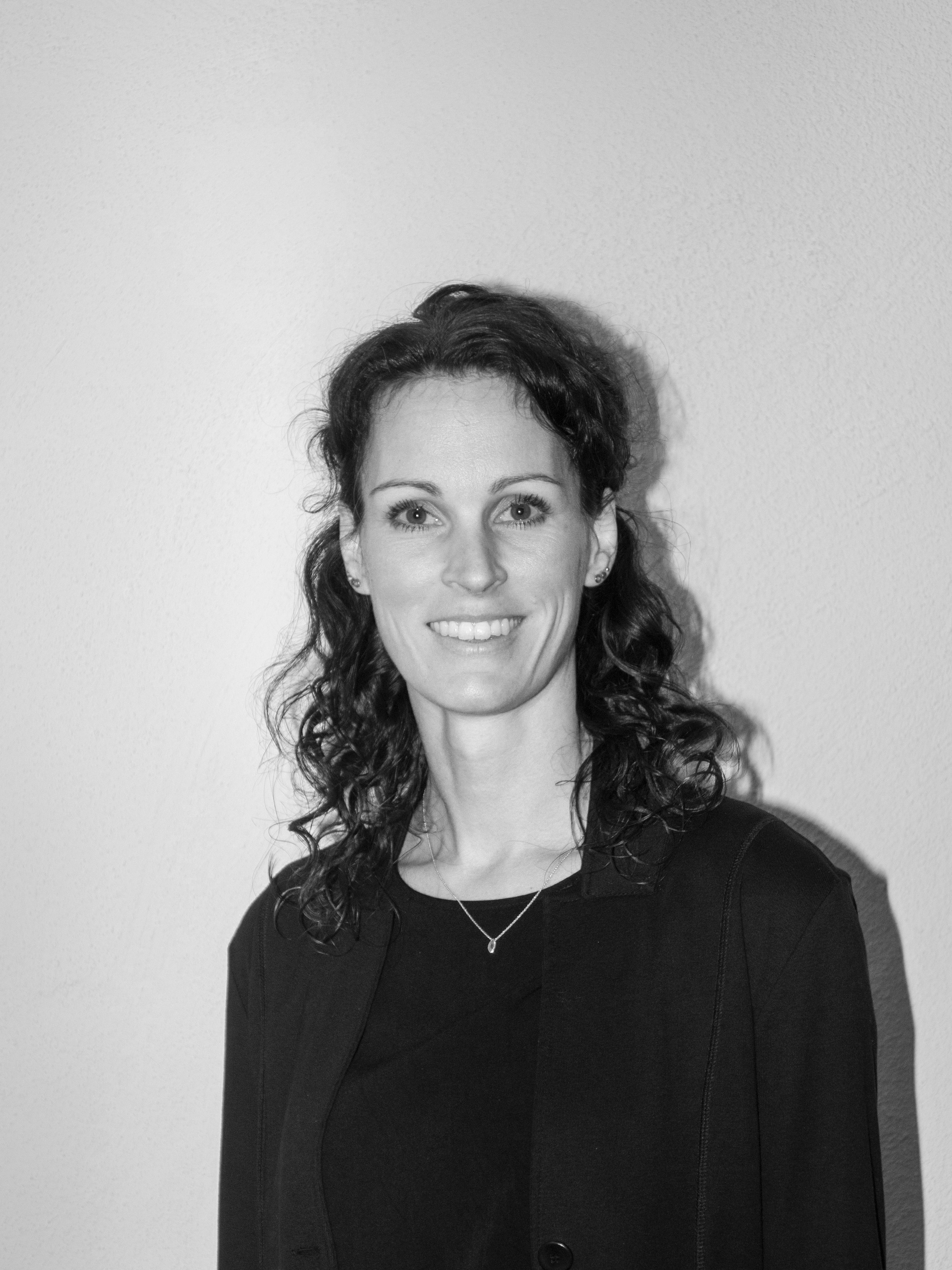 Schwarz-Weiß-Foto einer Frau mit lockigem Haar, die lächelt, vor einer einfarbigen Wand.