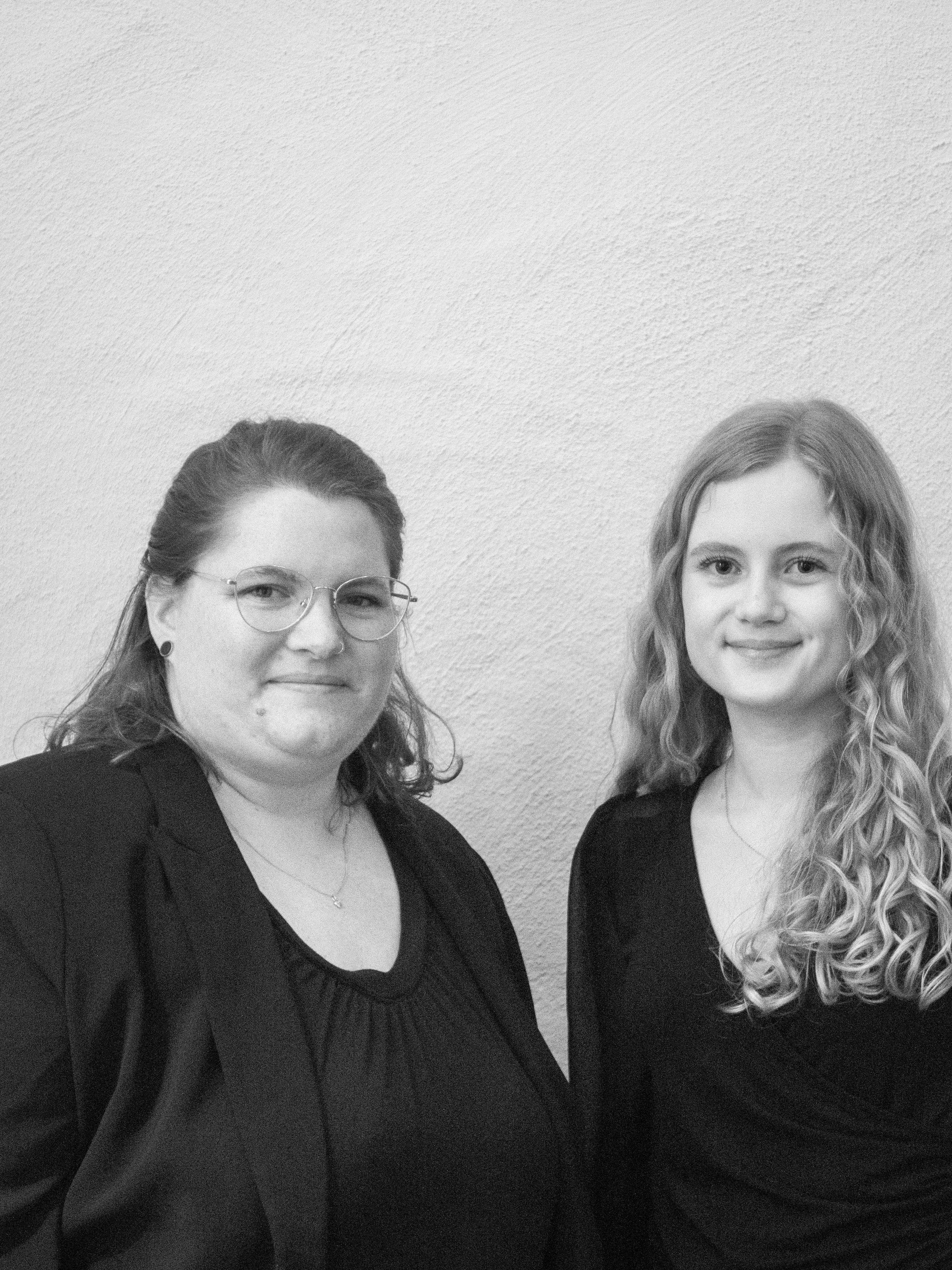 Zwei Personen in formeller Kleidung stehen vor einer hellen Wand. Eine Person trägt eine Brille und hat mittellange Haare, die andere Person hat lange gelockte Haare.