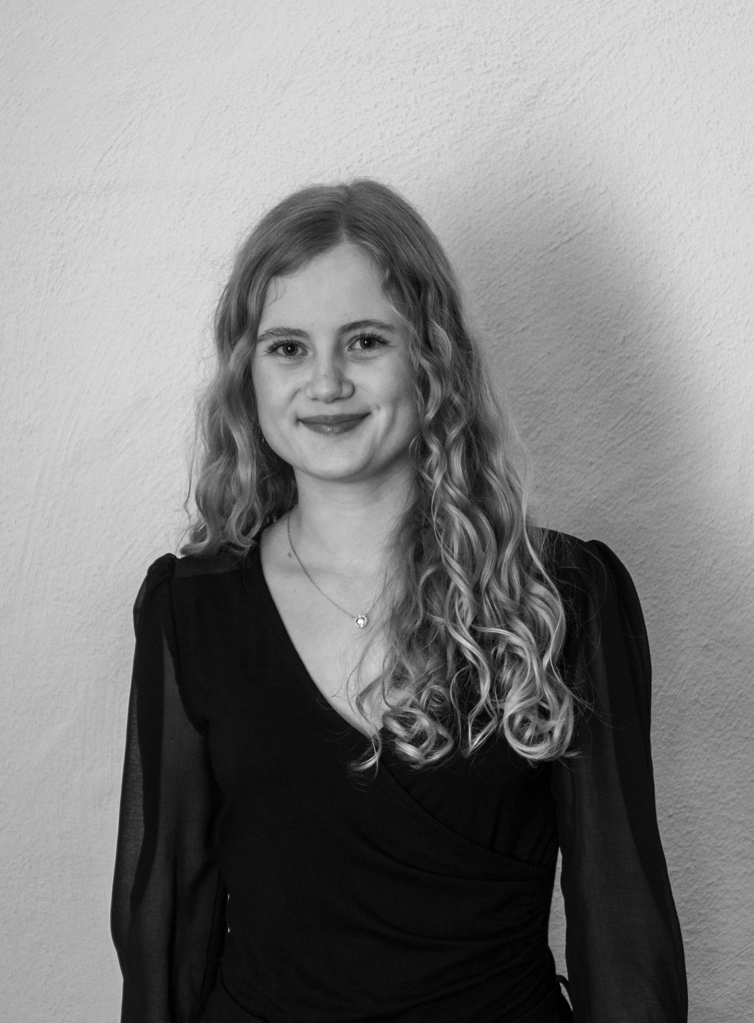 Schwarz-weiß Porträt einer lächelnden Frau mit langen, lockigen Haaren in einem schwarzen Kleid vor einem neutralen Hintergrund.