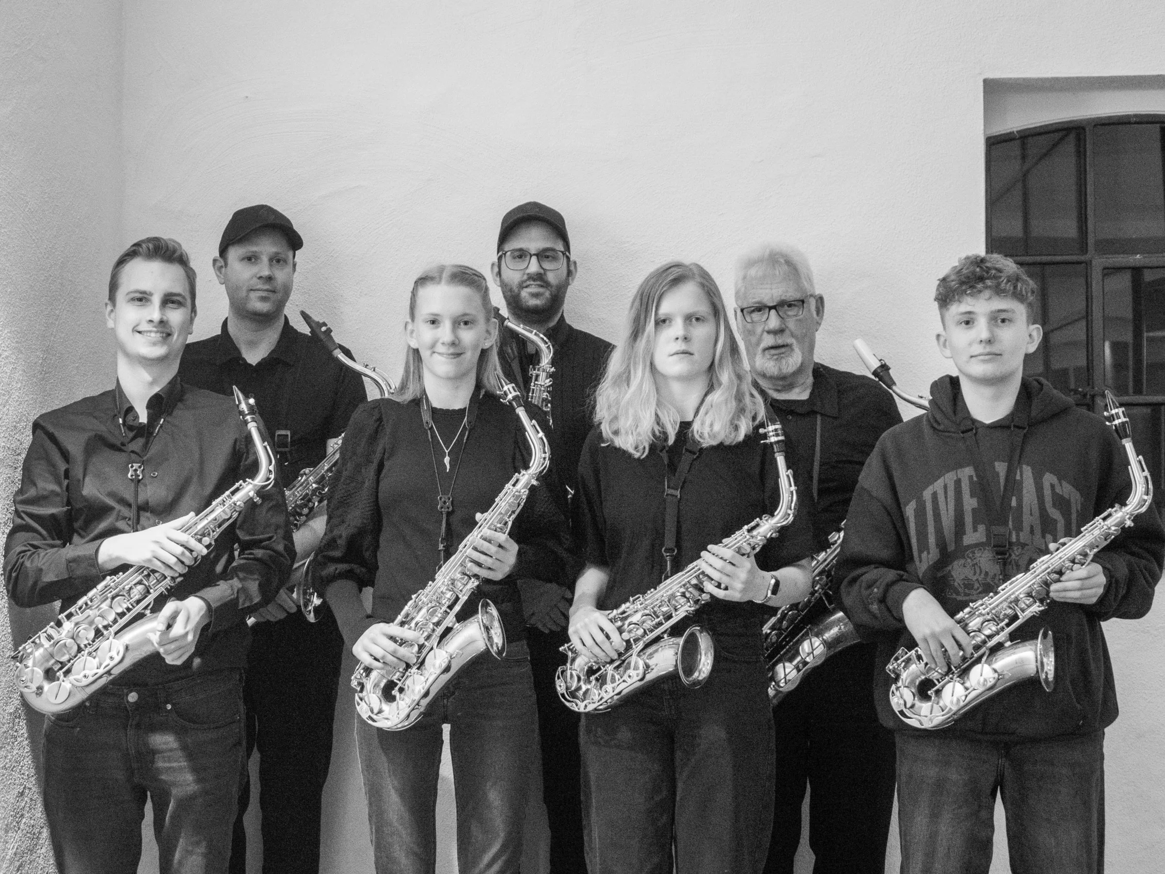 Eine Gruppe von sieben Personen, die Saxophone halten und in formeller Kleidung vor einer hellen Wand stehen.
