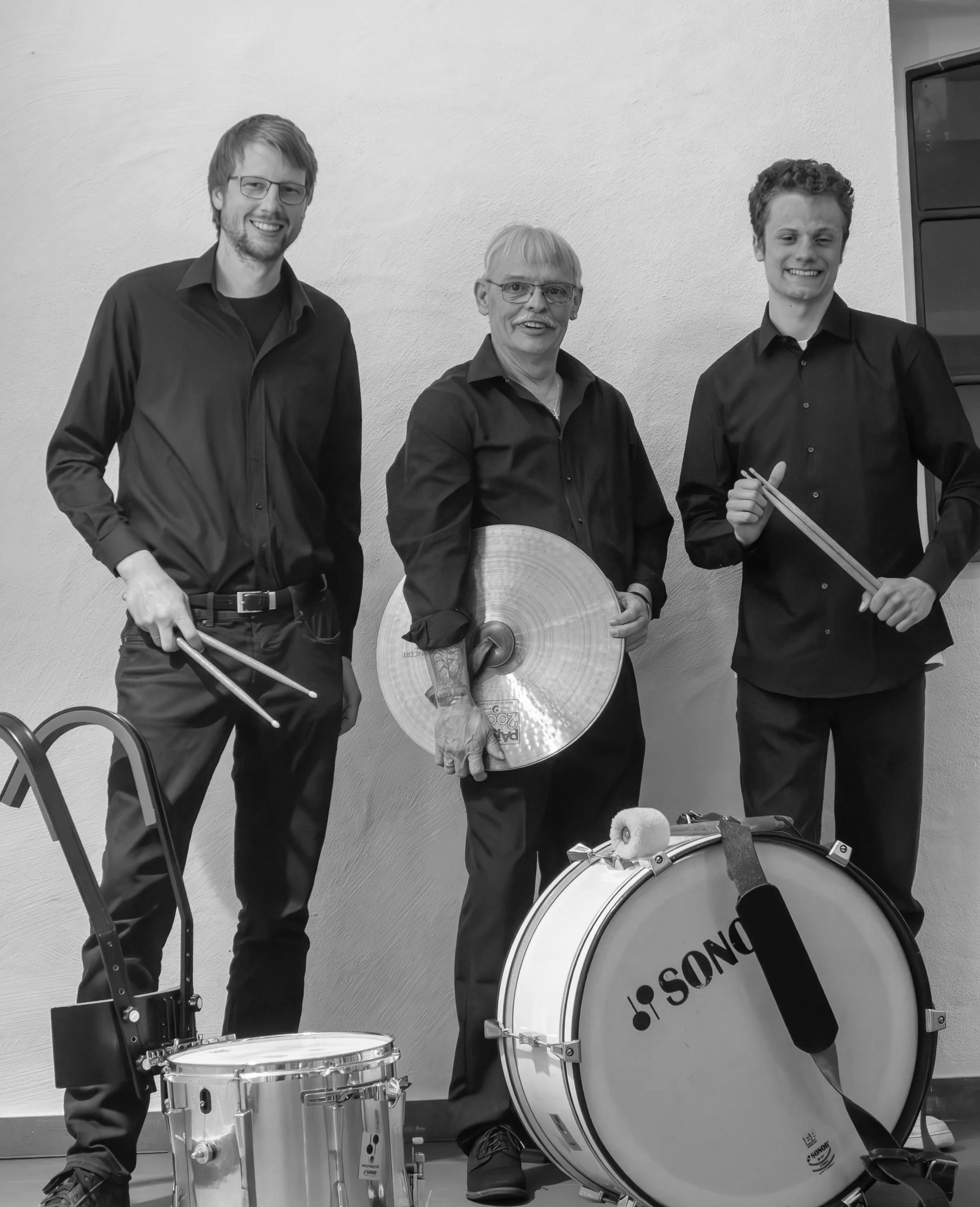 Drei Männer in schwarzer Kleidung mit Schlaginstrumenten posieren lächelnd vor einer weißen Wand. Einer hält Trommelstöcke, ein anderer ein Becken und der dritte steht neben einer großen Trommel mit der Aufschrift "SONOR".