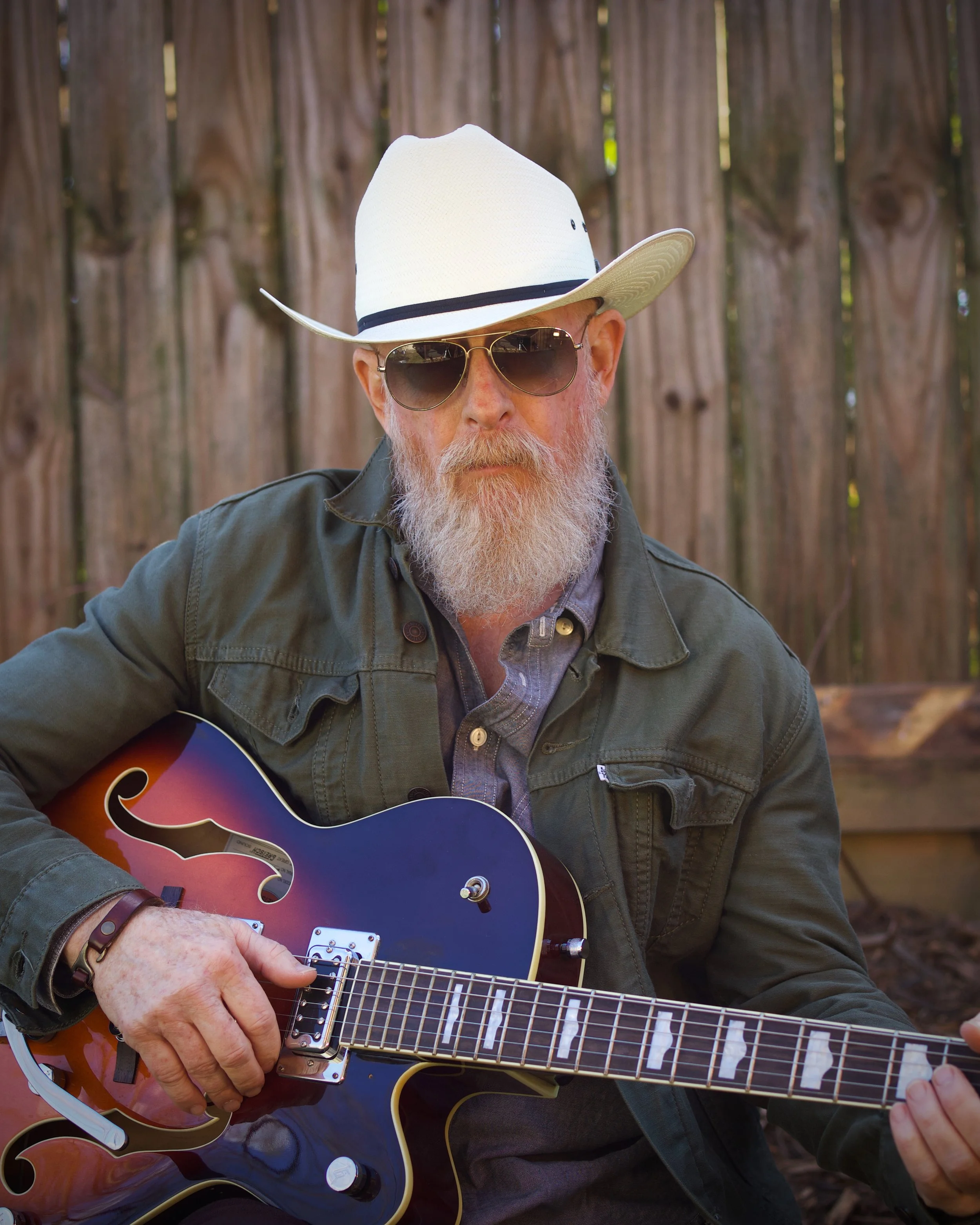 Old Hank with a white beard and sunglasses playing an electric guitar outdoors, wearing a white hat and a dark green jacket, with a wooden fence in the background.