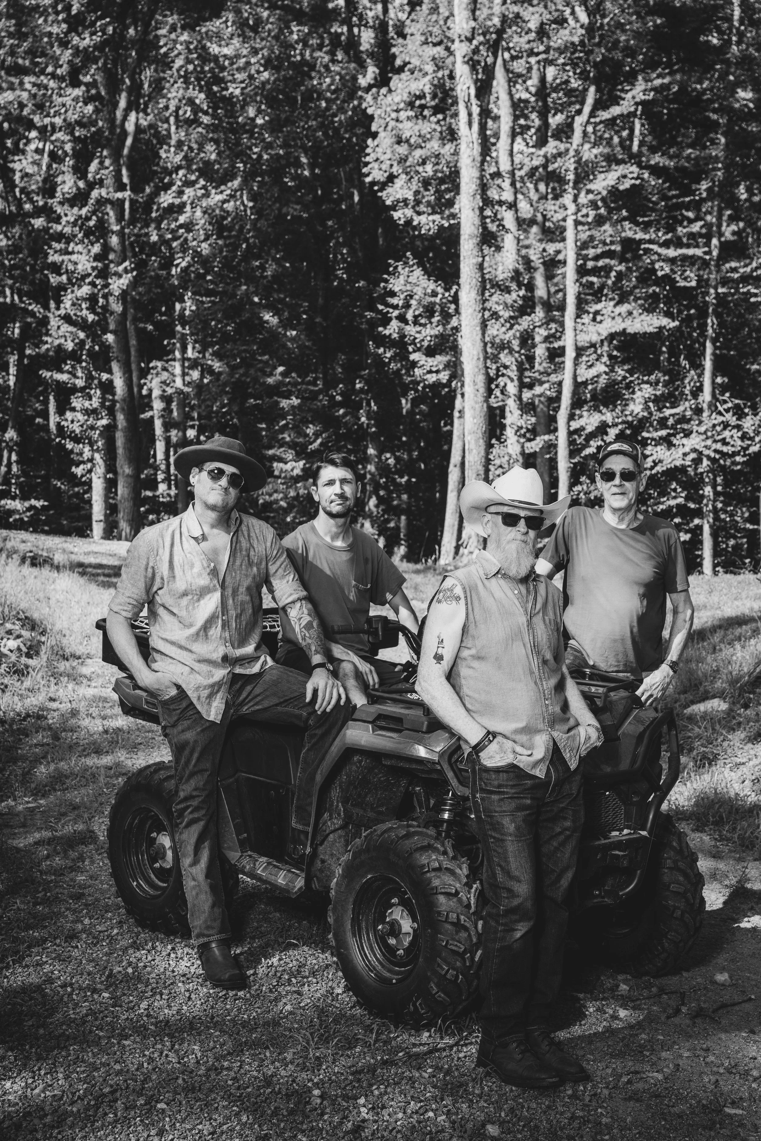 The 10-4 Good Buddies at Buck Naked Hunt Camp in casual clothing and sunglasses posing around and on a four-wheeler vehicle in a wooded outdoor area during daytime.