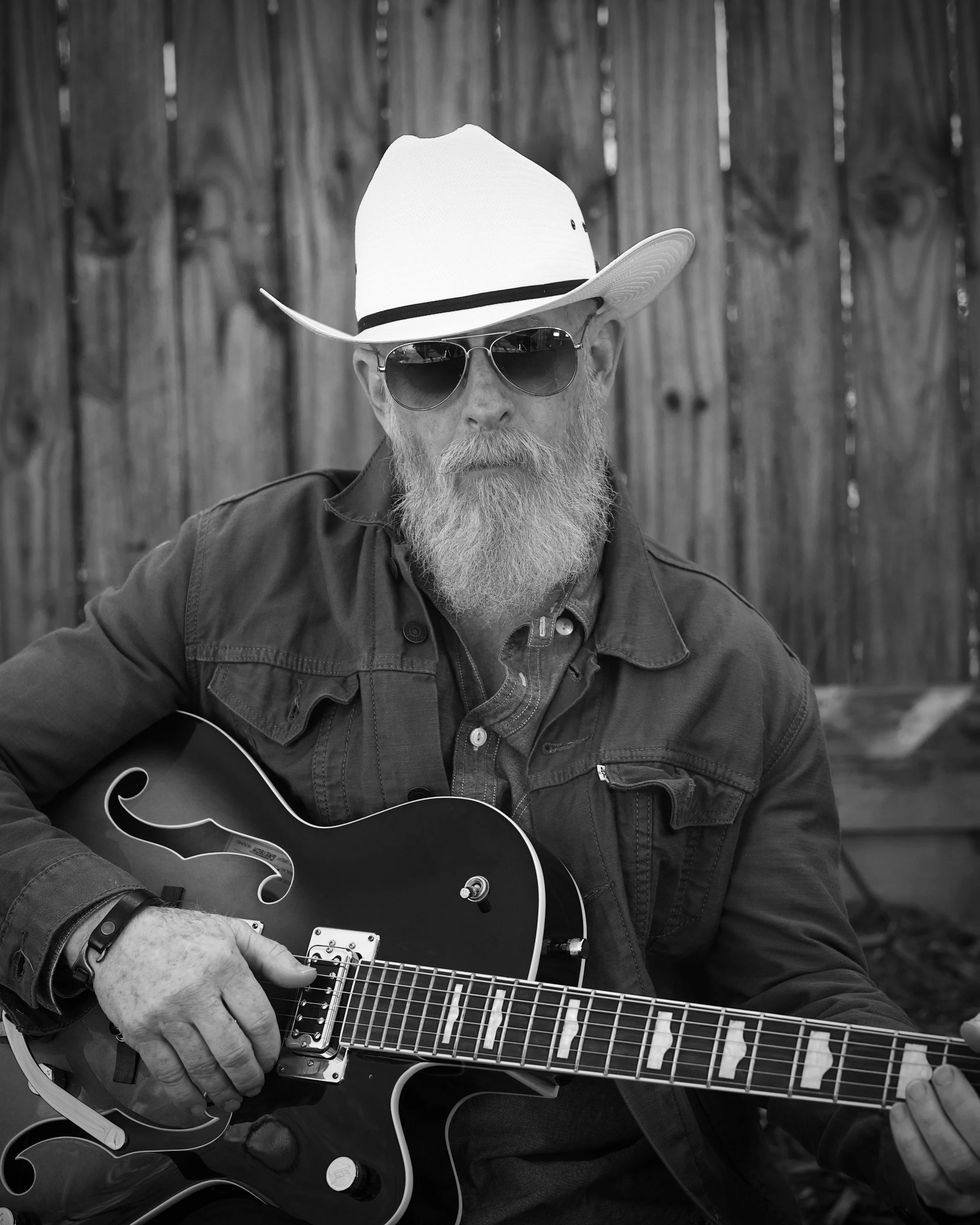 Old Hank with a beard wearing sunglasses, a hat, and a leather jacket, playing an acoustic guitar outdoors in front of a wooden fence.