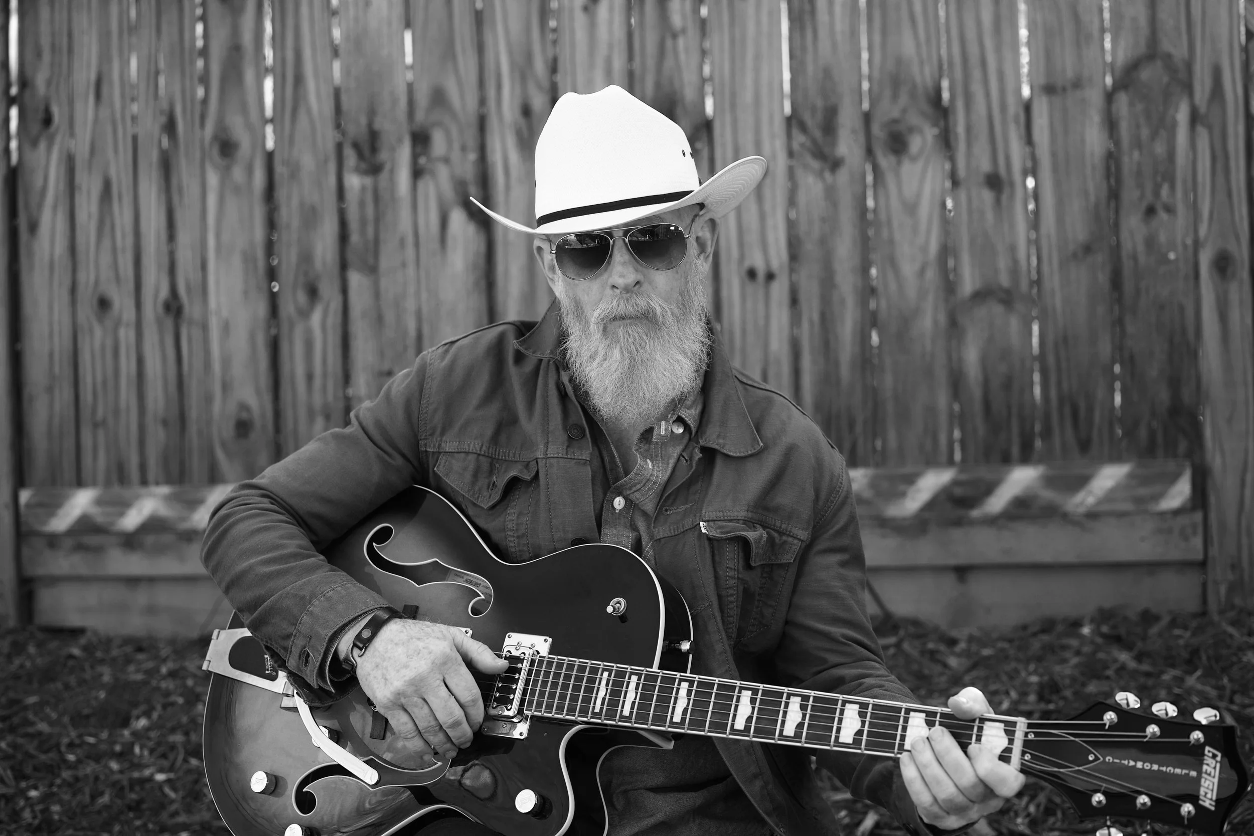 Old Hank with a beard and sunglasses, wearing a cowboy hat, playing an electric guitar outdoors in front of a wooden fence.