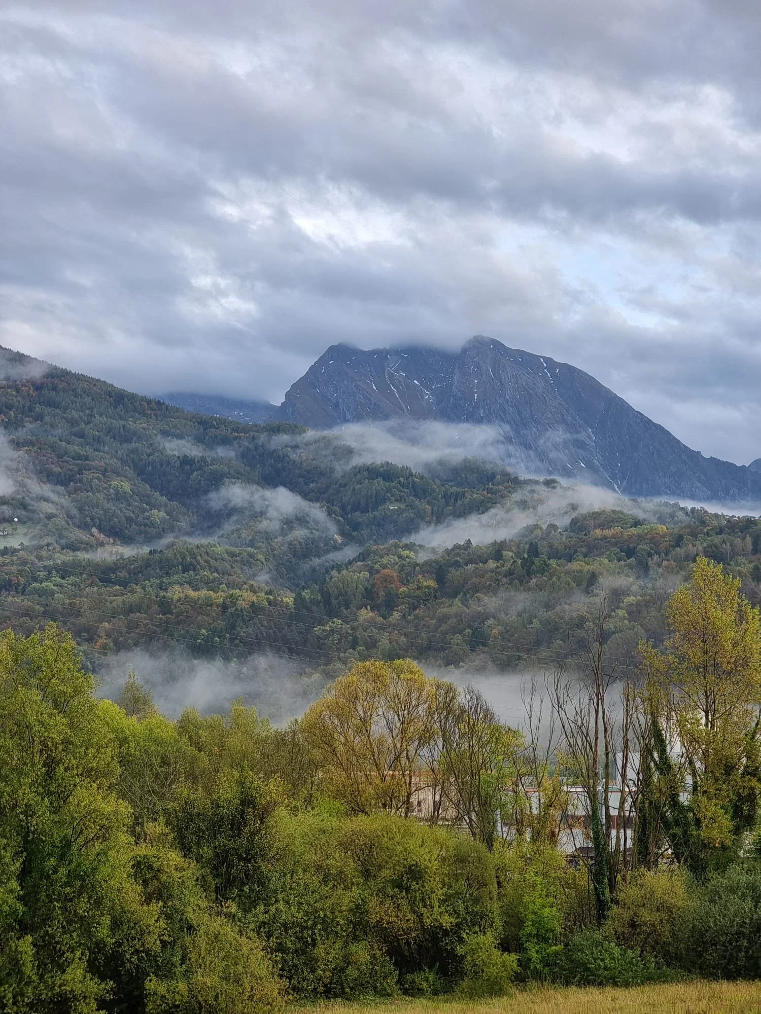 Paesaggio montano con montagne alte e coperta di nuvole, boschi verdi e foschie sulla valle.