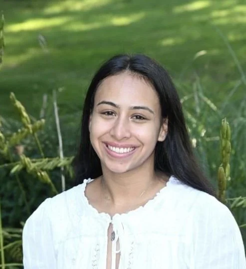A woman with long dark hair smiling in a green outdoor setting with tall plants.