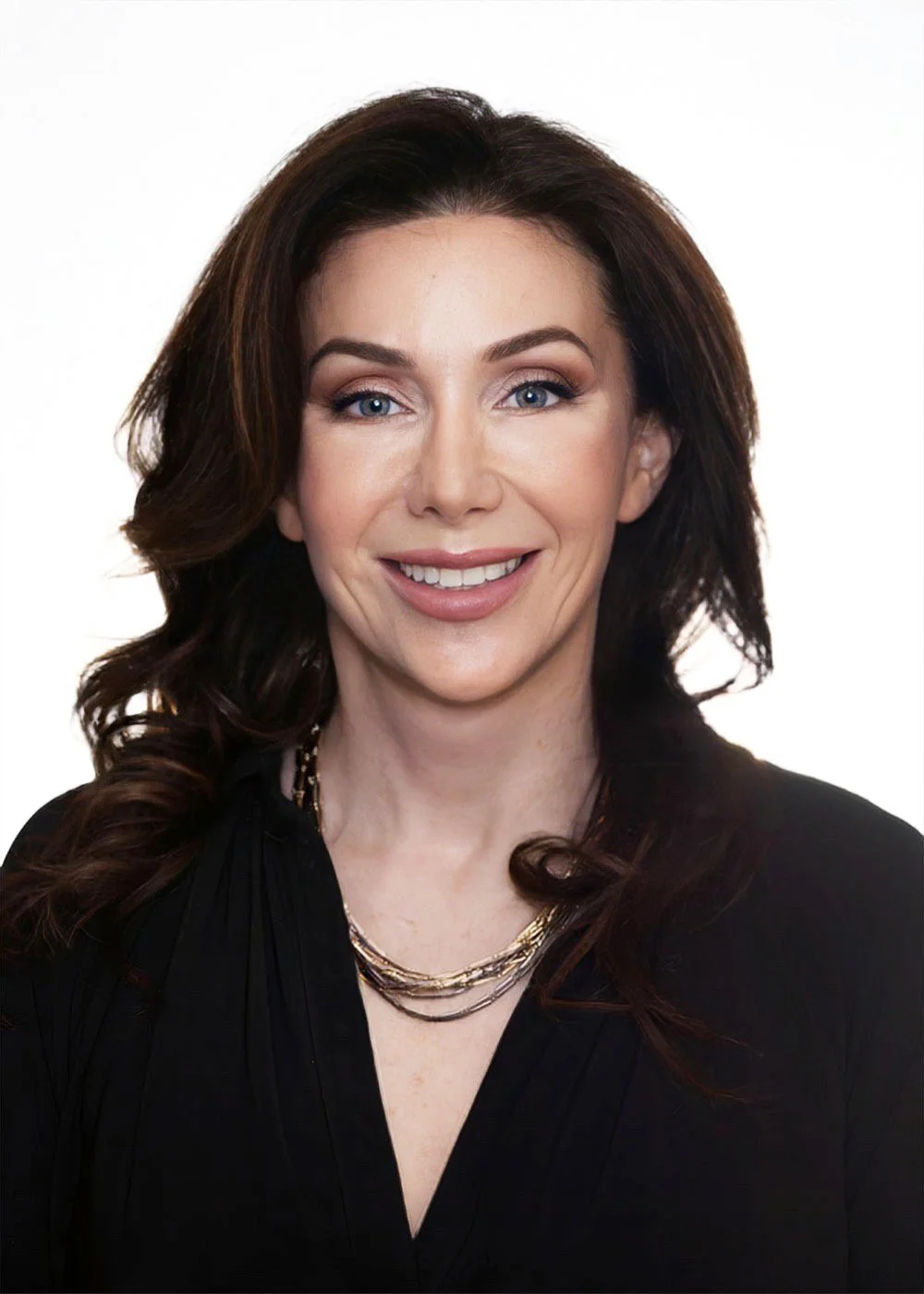 A woman with long dark wavy hair, blue eyes, and fair skin, smiling, wearing a black top with layered gold necklaces against a white background.