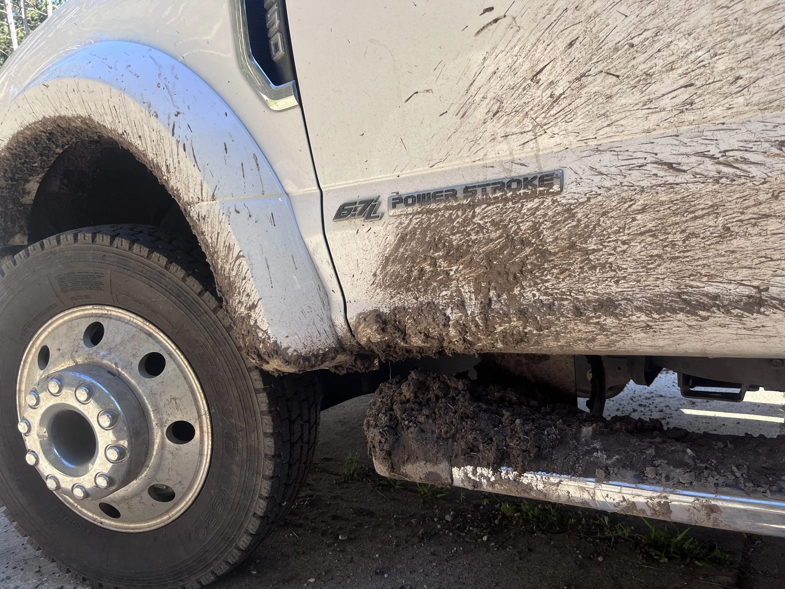Close-up of a dirty white pickup truck with mud splattered on the side, showing a badge that reads '6.7L Power Stroke' and a muddy tire.