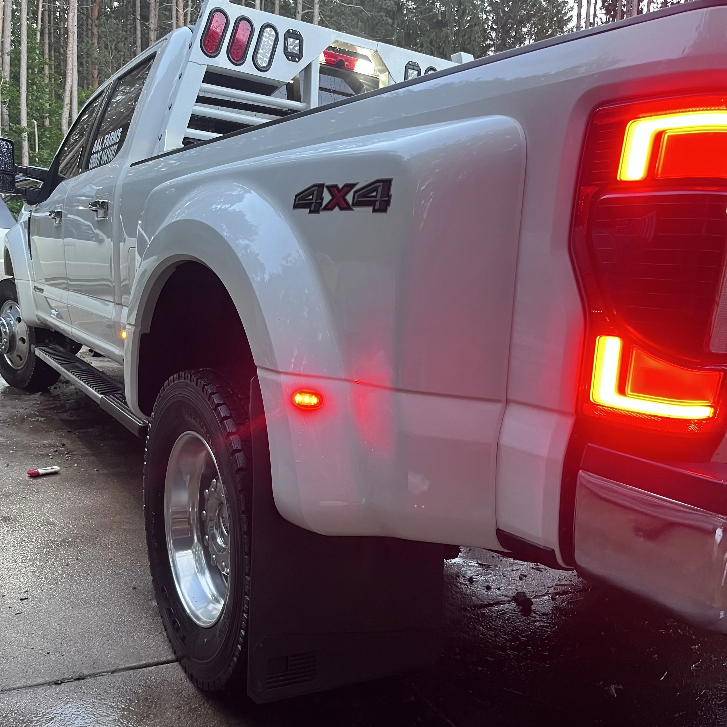 Close-up of the rear right side of a white 4x4 pickup truck, showing tail lights, wheel, and a reflective red side marker, parked on a wet road.