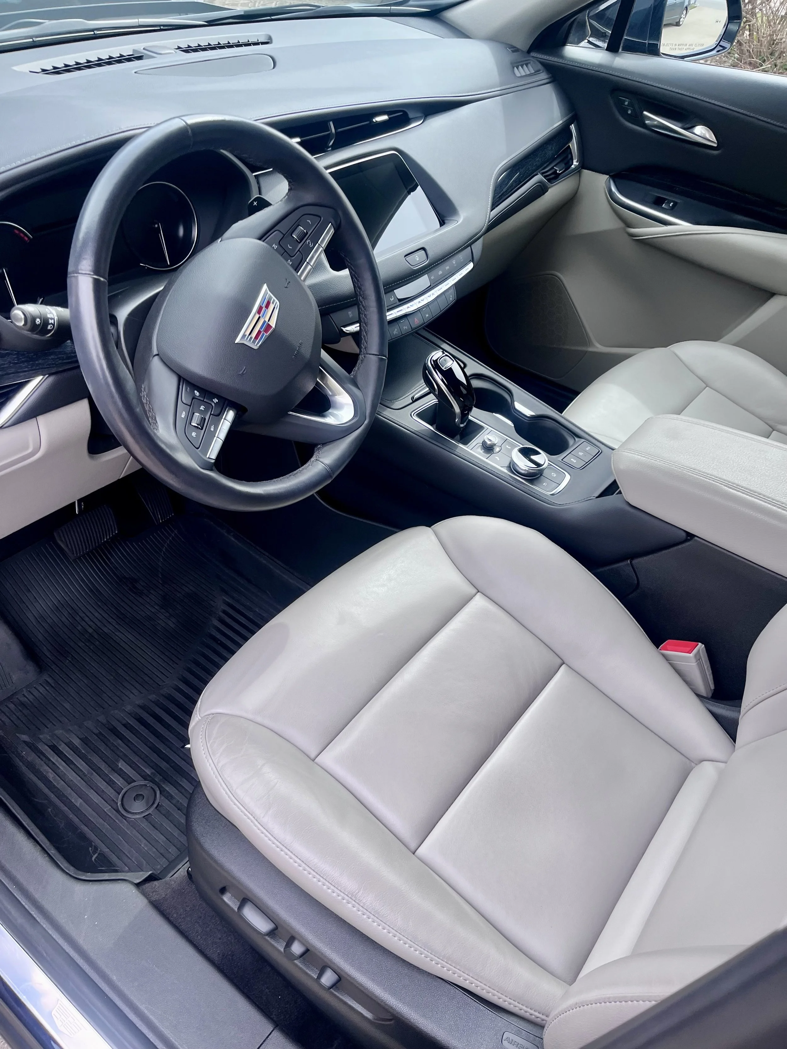 Interior of a modern Cadillac vehicle, showing a black leather steering wheel with a Cadillac emblem, a digital dashboard, a touchscreen display, gear shift, and beige leather front seats.