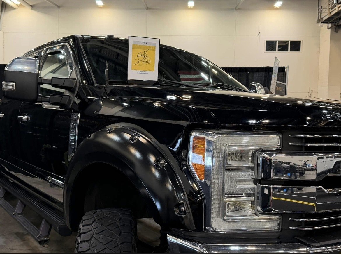 Side view of a black pickup truck with large tires, chrome grille, and mirror, displayed at an indoor car show with signs on the windshield.