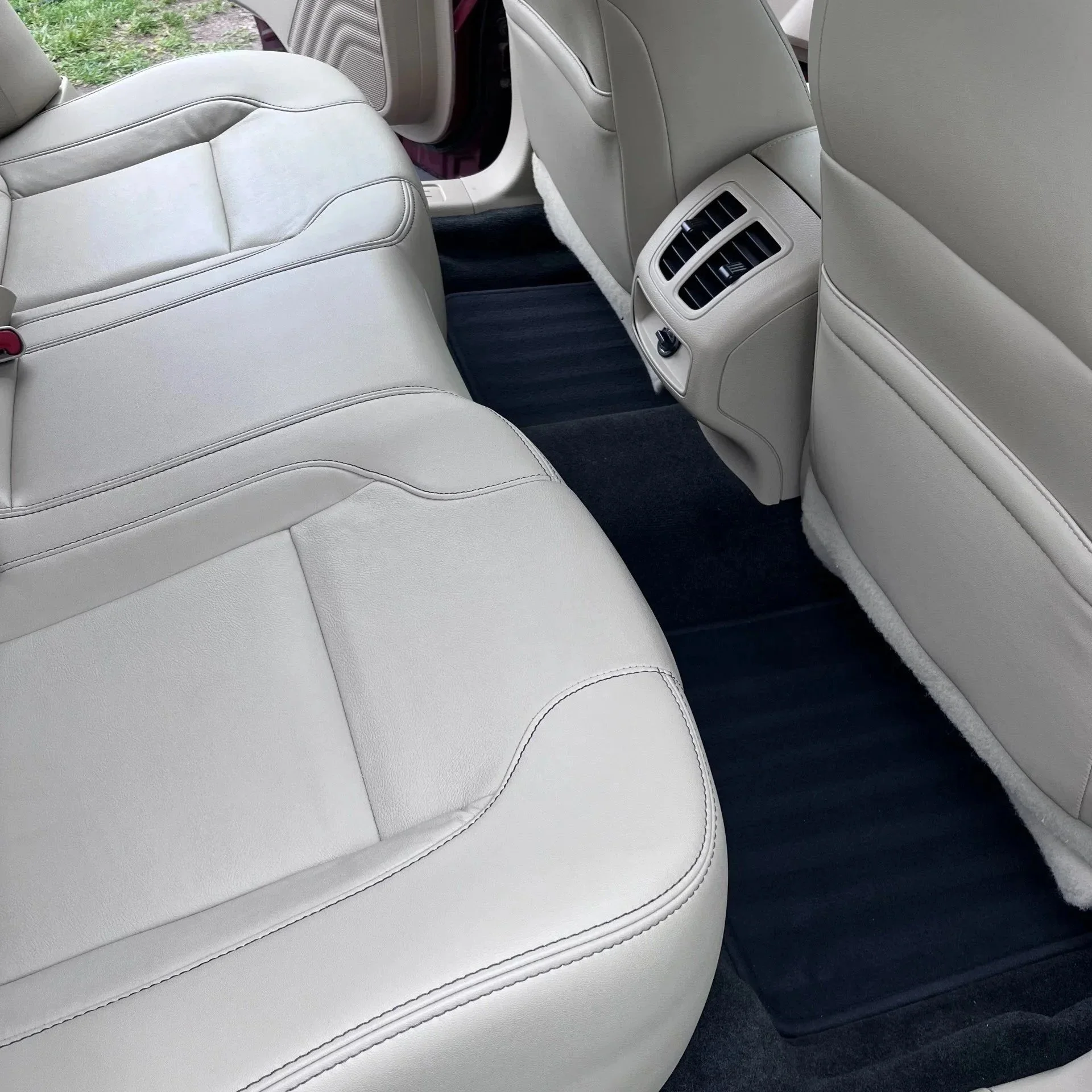 The backseat interior of a car with light-colored leather seats, air vents, and black floor mats.