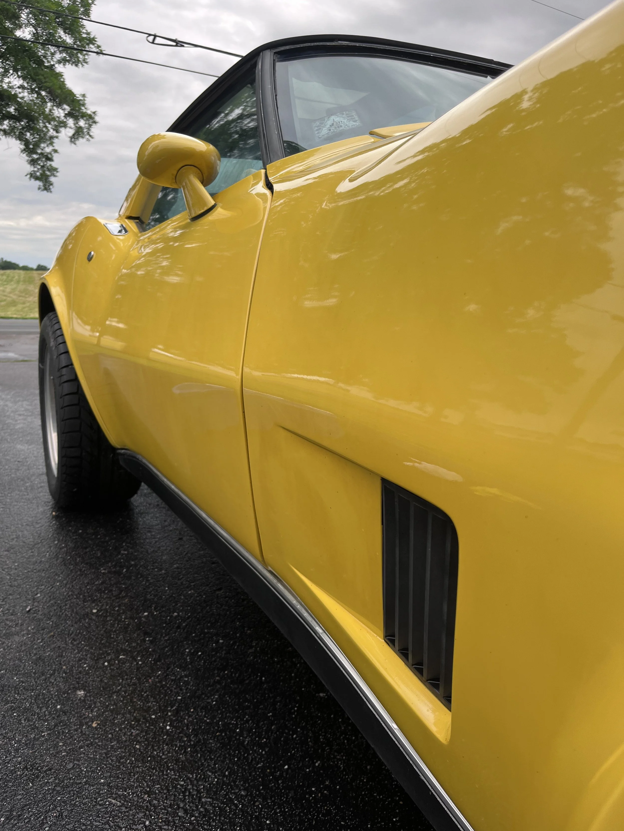 Close-up of a yellow vintage sports car, showing part of the front side, mirror, door, and a black vent on the side panel, parked on a wet asphalt road with trees and cloudy sky in the background.
