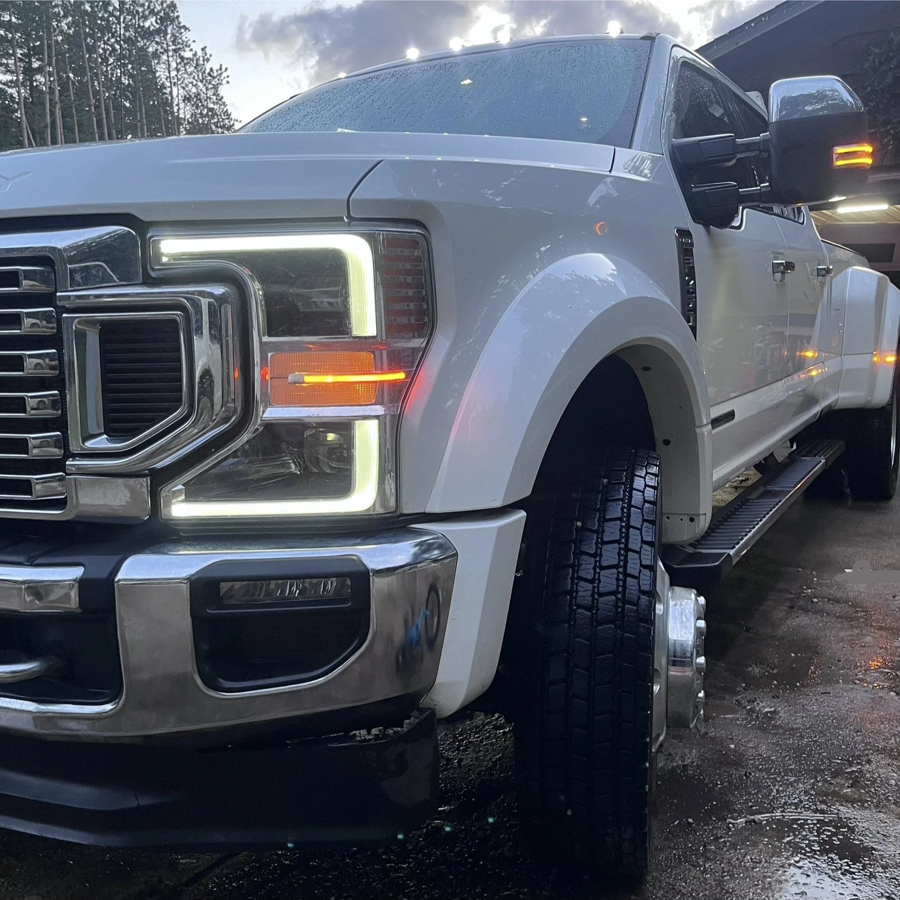 Close-up of a white heavy-duty truck with a large front grille and bright LED headlights parked outdoors on a wet surface, with trees and sky in the background.