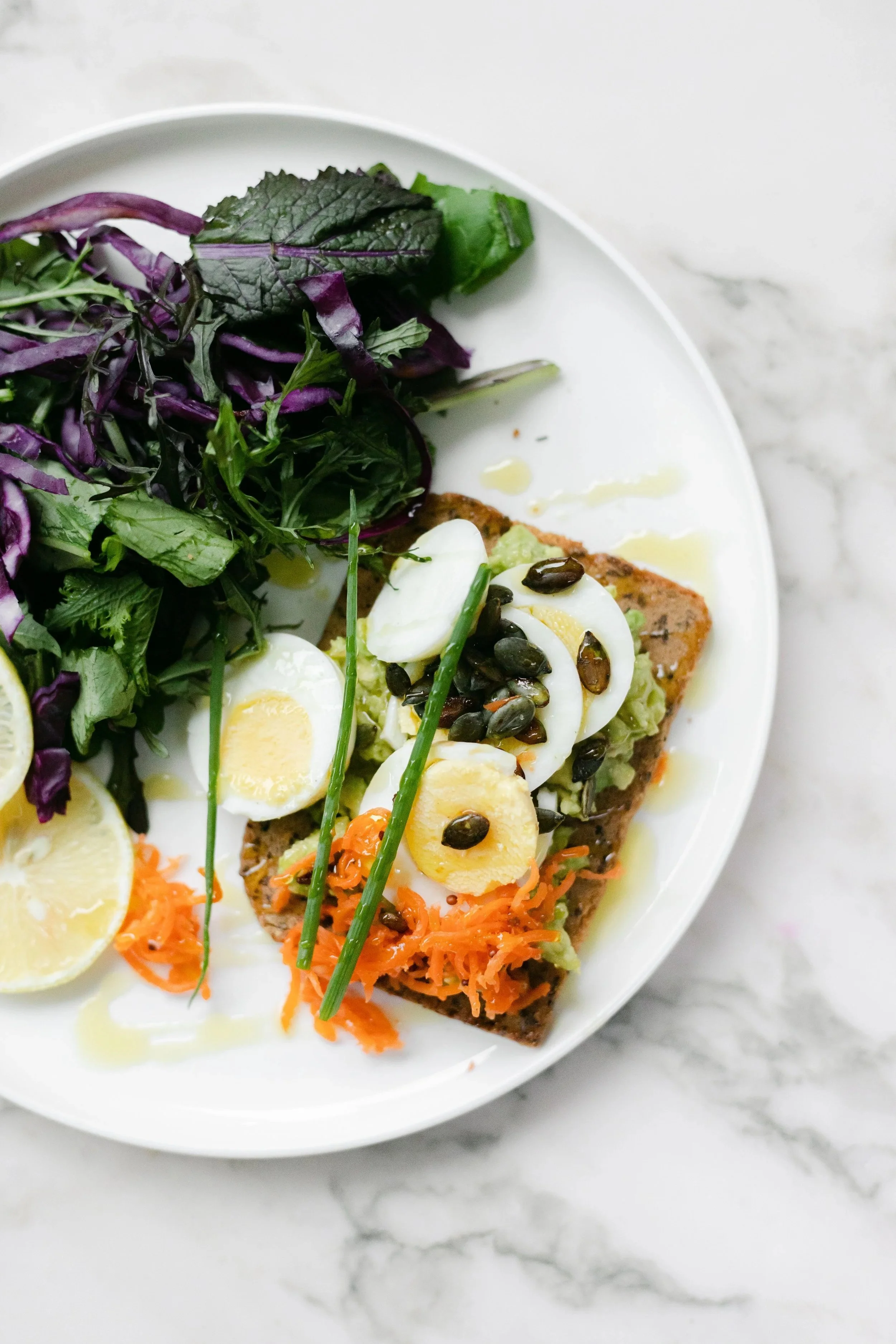 A white plate with salad greens, sliced boiled eggs topped with sunflower seeds, and a piece of toasted bread with shredded carrots and chopped greens on top, drizzled with olive oil on a marble surface.