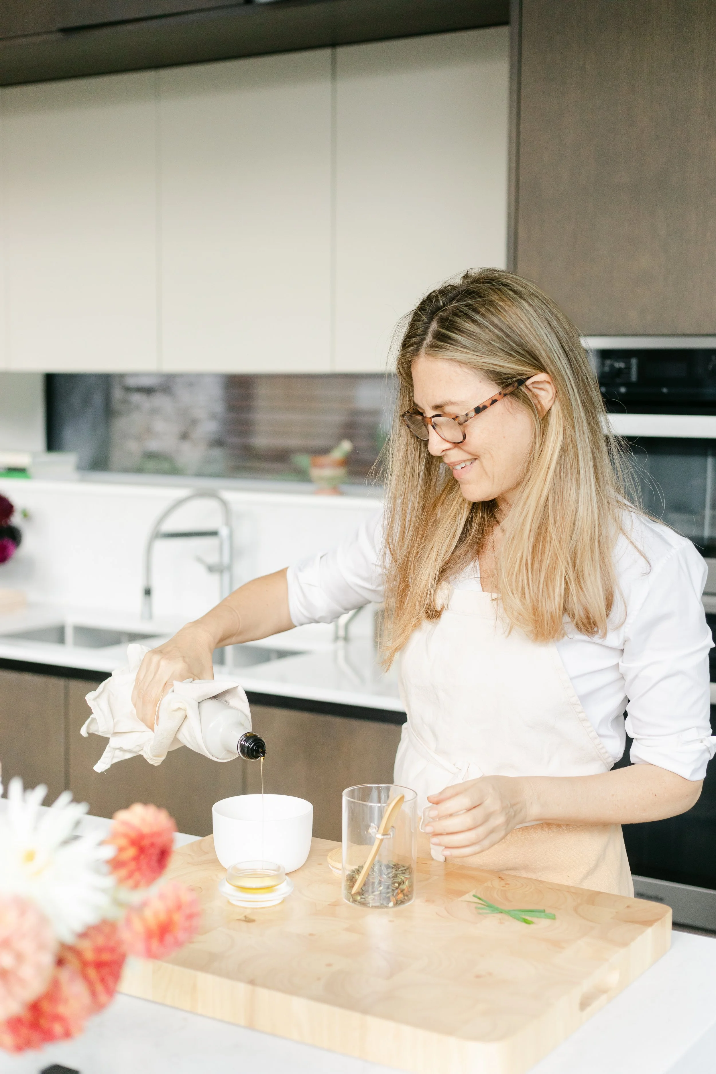 Woman with long blonde hair and glasses, wearing a beige apron, pouring liquid into a white cup on a kitchen countertop.