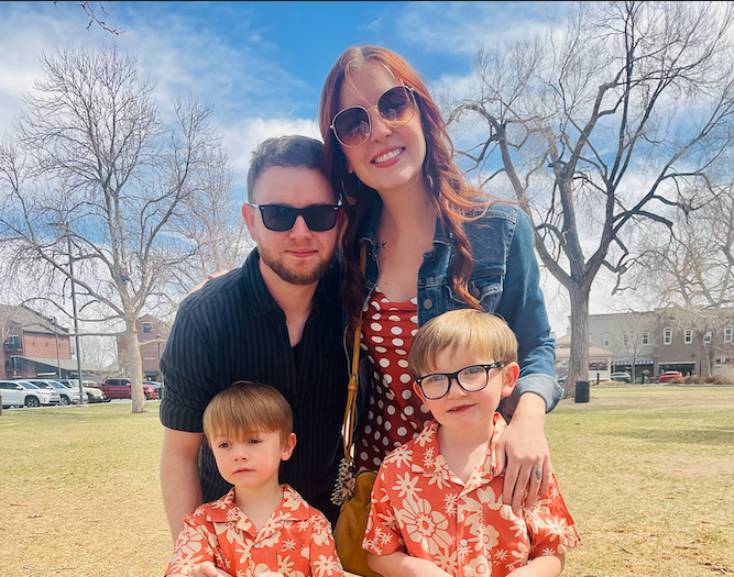 A family of four outdoors on a sunny day, with two adults and two children. The woman has long red hair, sunglasses, and a denim jacket. The man has dark hair, sunglasses, and a black shirt. The children have light brown hair and wear matching orange Hawaiian shirts. The background features leafless trees, a grassy field, and buildings.