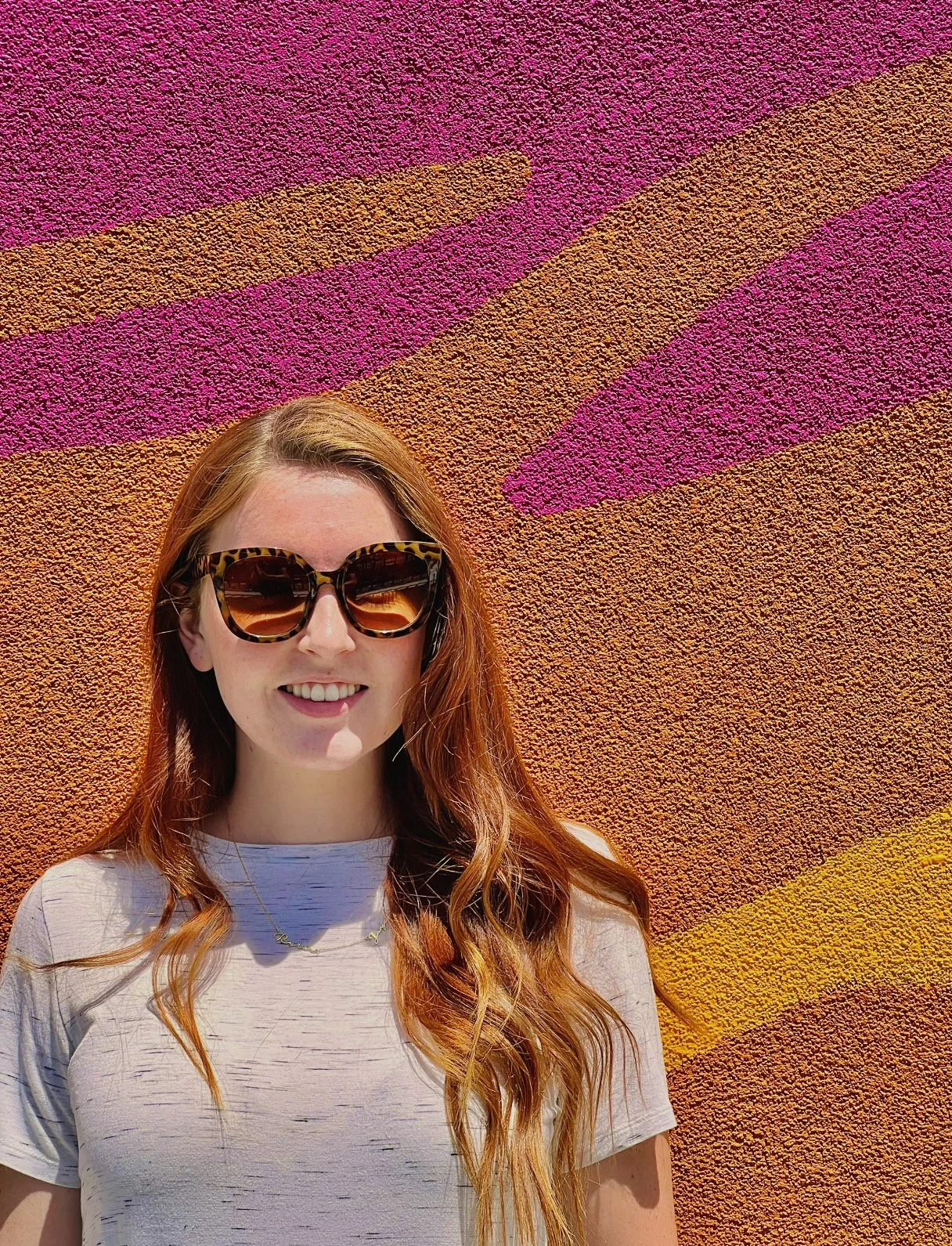 A woman with long red hair wearing sunglasses and a light-colored t-shirt standing in front of a colorful, textured wall with pink, orange, purple, and yellow patterns.