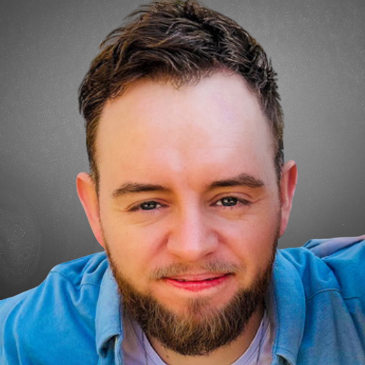 A man with a beard and brown hair smiling at the camera wearing a blue shirt, gray background.