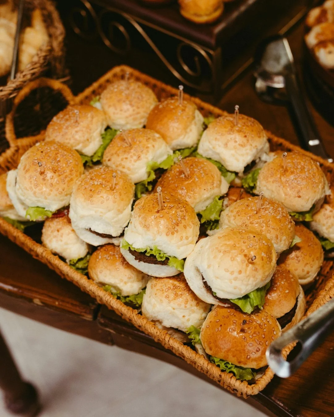 Mini cheeseburgers with lettuce, tomato, cheese, bacon, and sesame seed buns on a white rectangular plate.