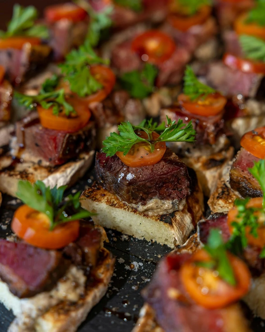 Person holding a broken piece of chocolate cookie with chocolate chips, with more cookies on a sheet of parchment paper in the background.