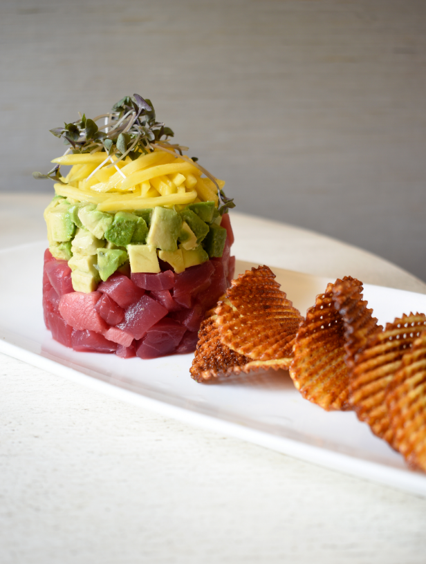 A tiered stack of diced raw tuna, avocado, and mango, garnished with microgreens, served with ridged potato chips on a white plate.