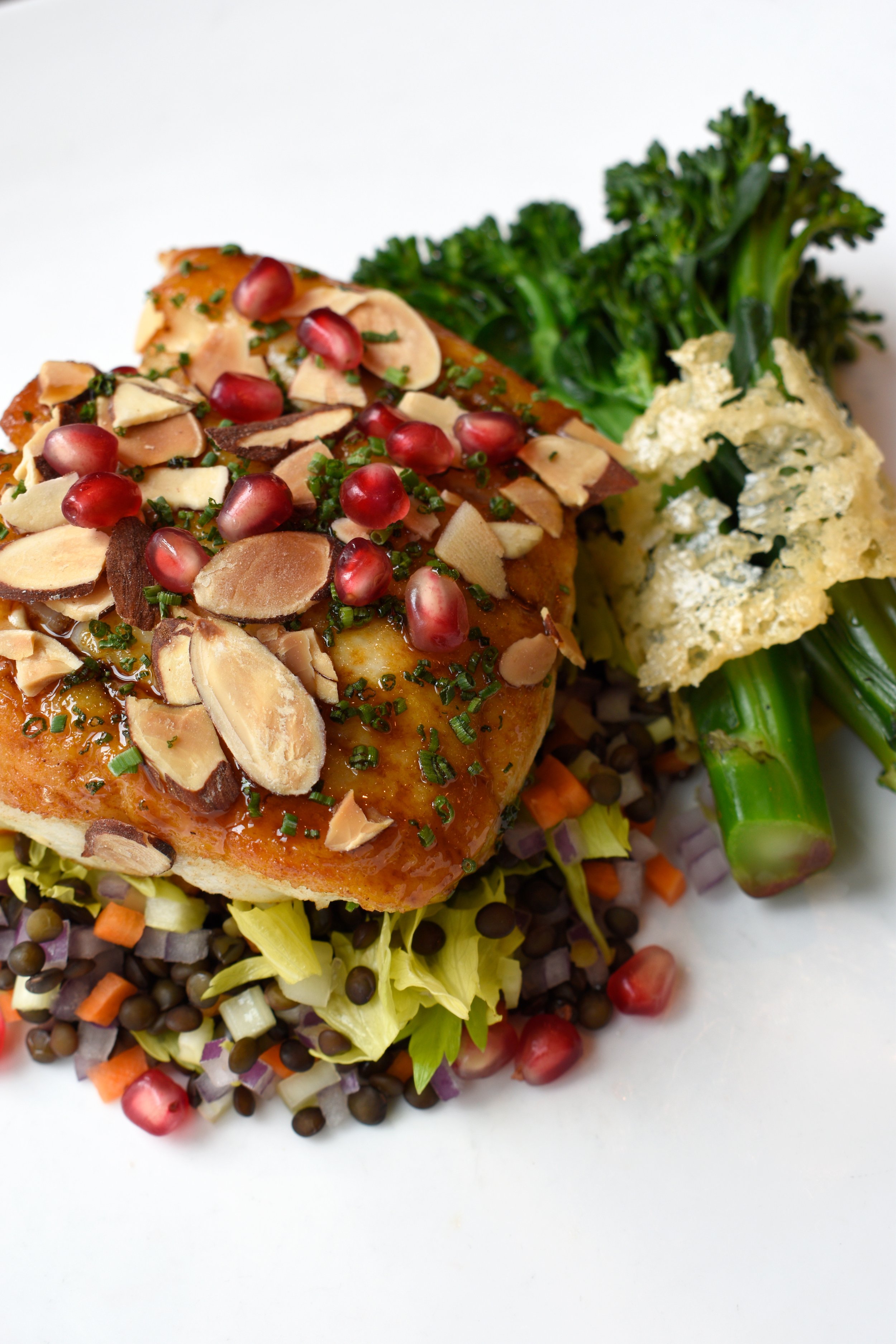 A plated dish with a piece of cooked fish topped with sliced almonds and pomegranate seeds, served on a bed of chopped vegetables with broccolini and crispy fried kale.