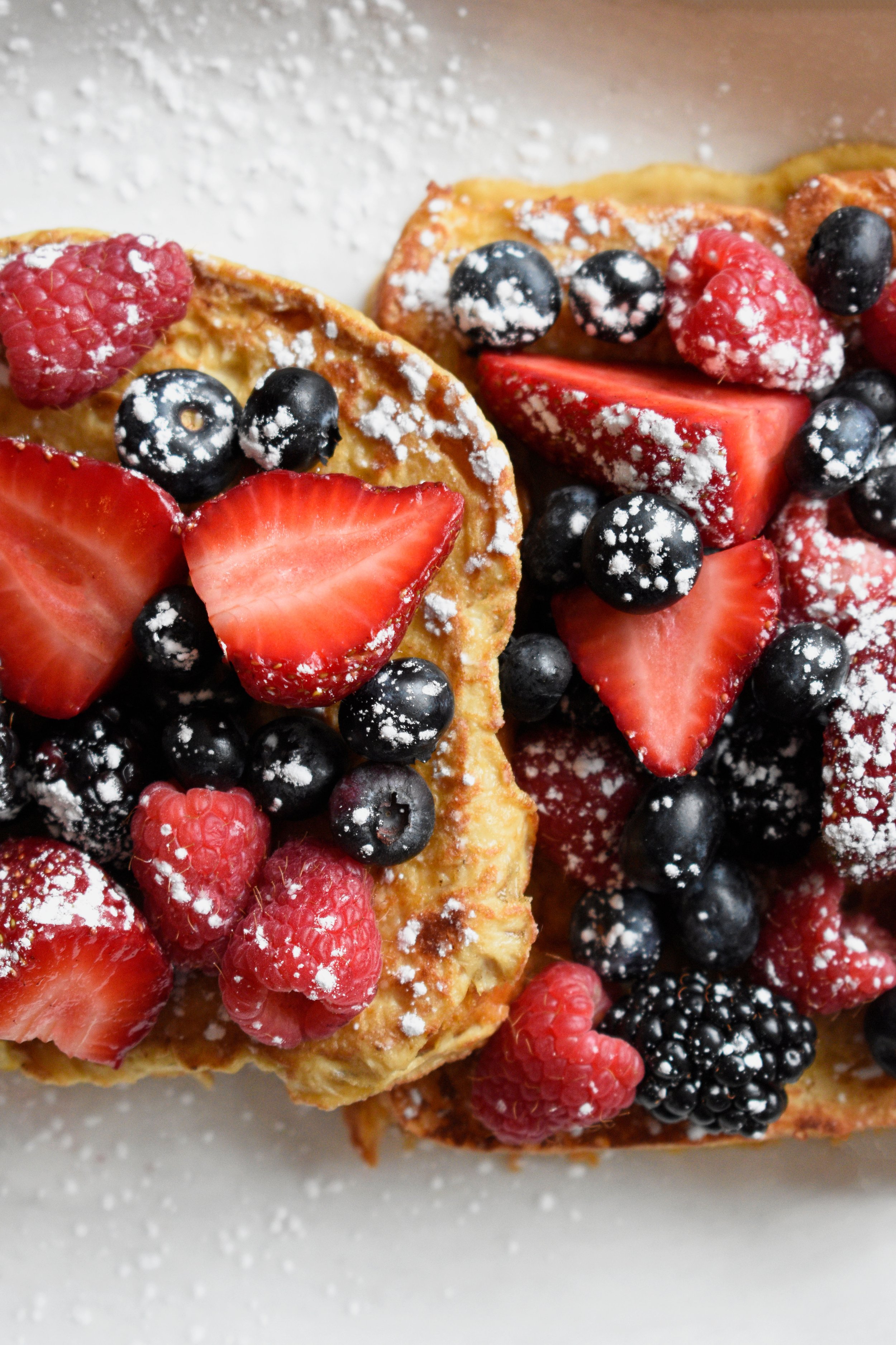 Close-up of two slices of French toast topped with strawberries, blueberries, raspberries, and powdered sugar.