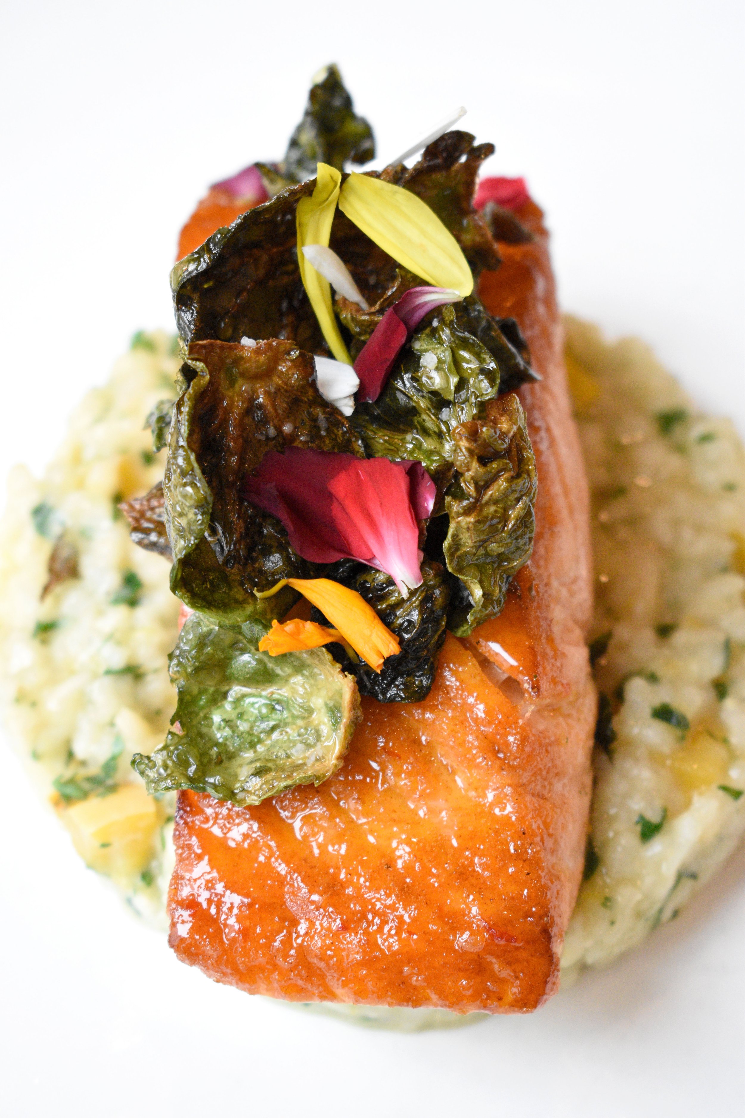 A plated dish featuring a cooked salmon fillet topped with crispy seaweed and edible flower petals, served alongside a creamy parsley potato mash.