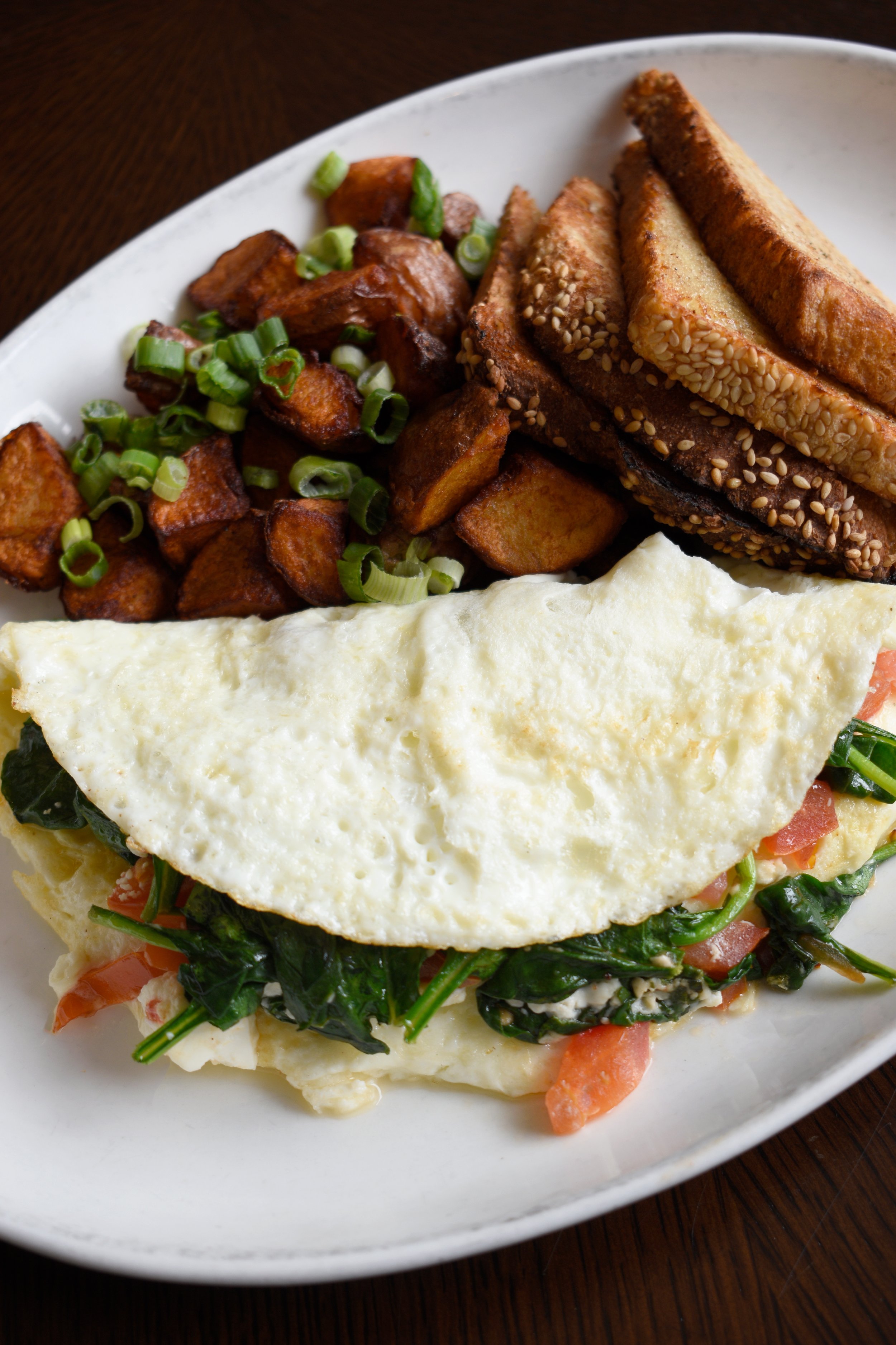 Breakfast plate with fried potatoes garnished with green onions, scrambled eggs with tomatoes and spinach, toasted bagel slices topped with sesame seeds, and scrambled eggs.