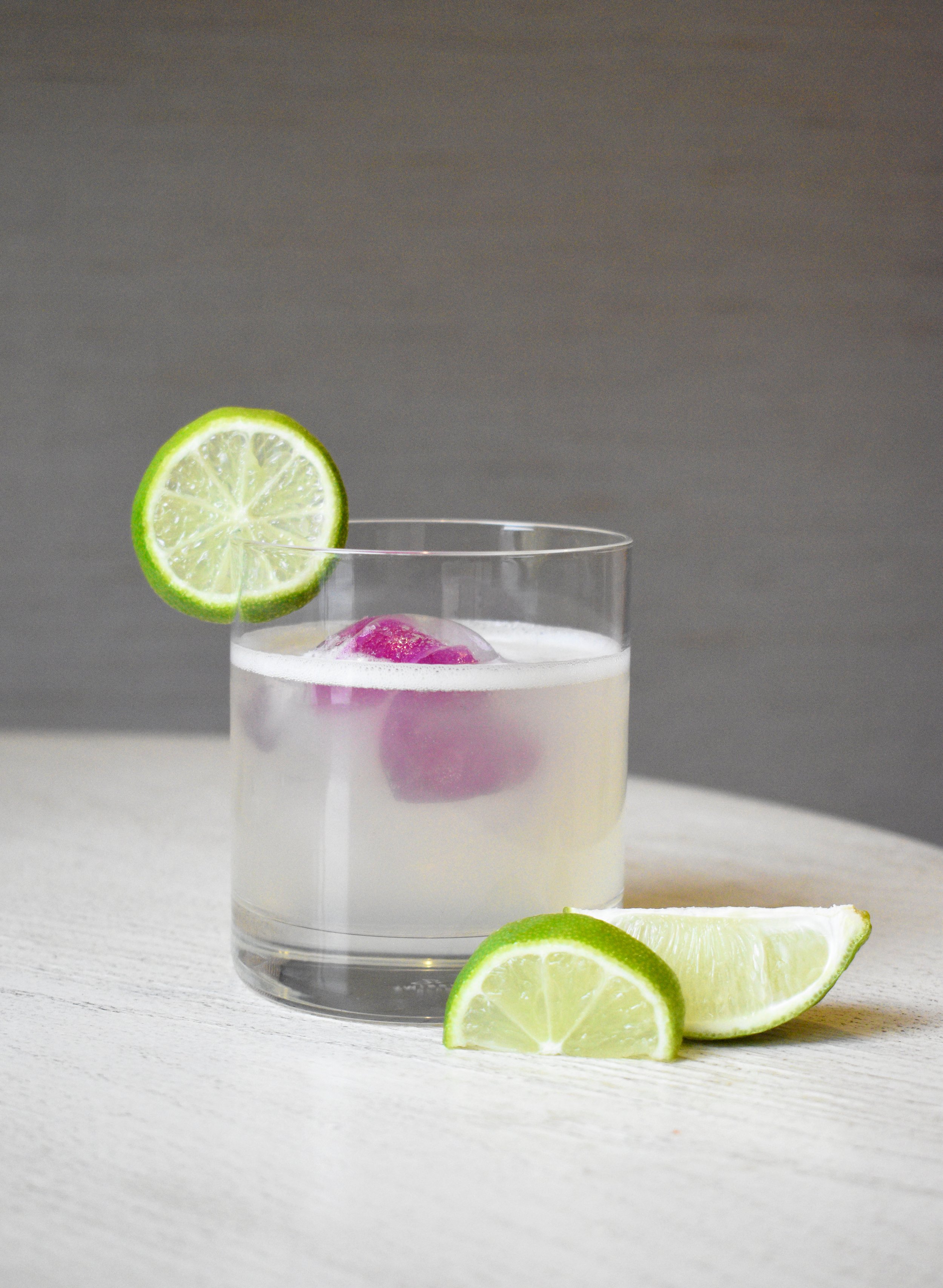 A glass of clear cocktail with ice, garnished with a lemon-lime slice on the rim and lime wedges on the table.