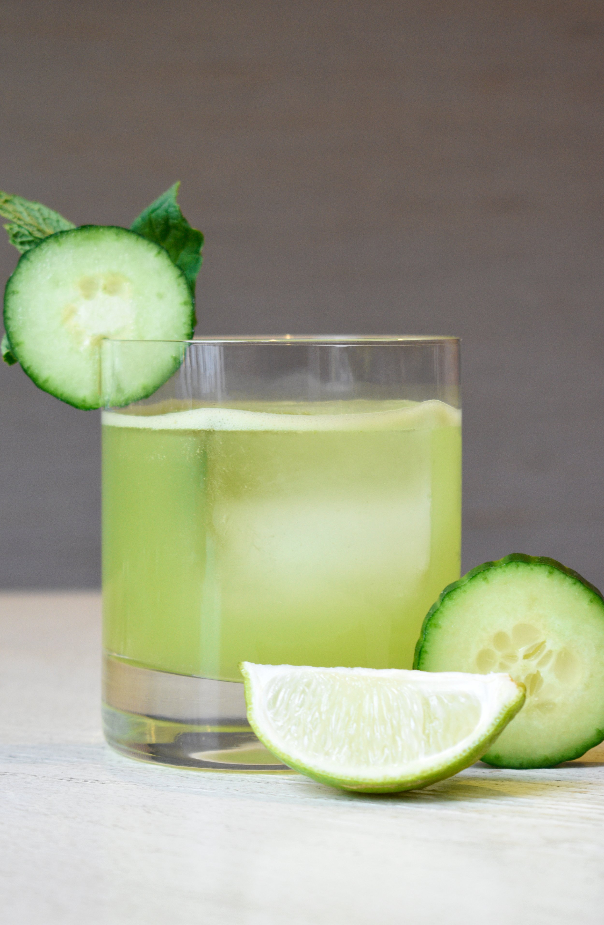 A glass of cucumber lime cocktail garnished with a slice of cucumber and a lime wedge on a light surface.