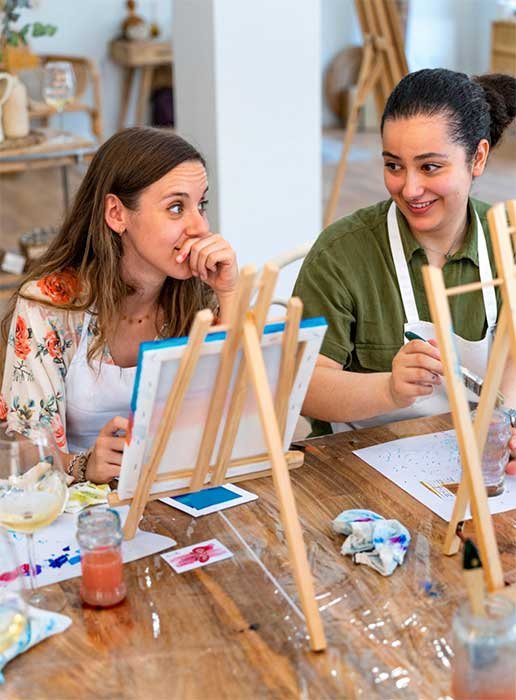 Two women at a paint-and-sip wine event,  sit at a table in front of table-top easels, painting on stretched canvases.