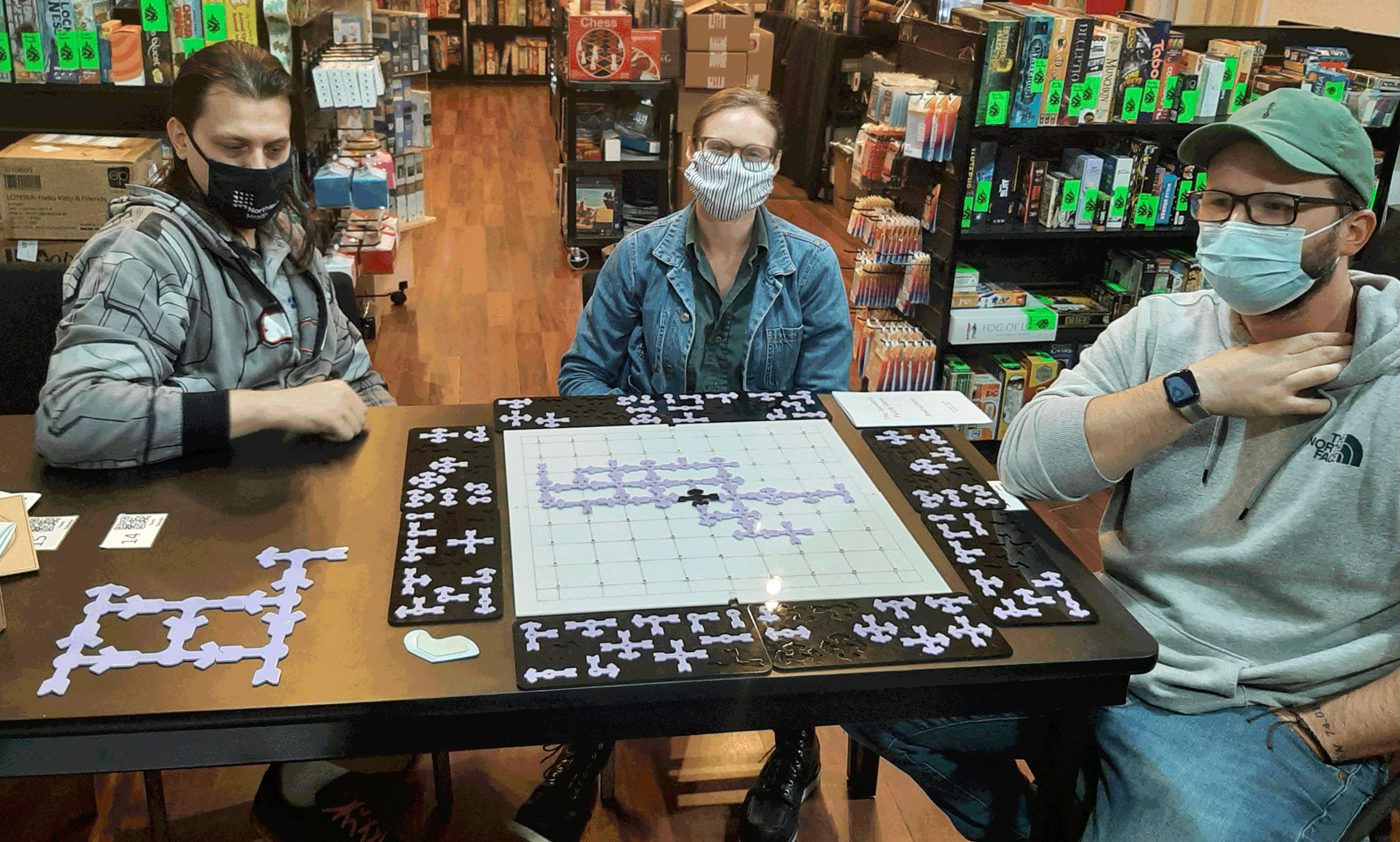 Three people, during Covid wearing masks, are sitting around a table playing a board game in a bookstore. The game involves assembling purple puzzle pieces on a the game board