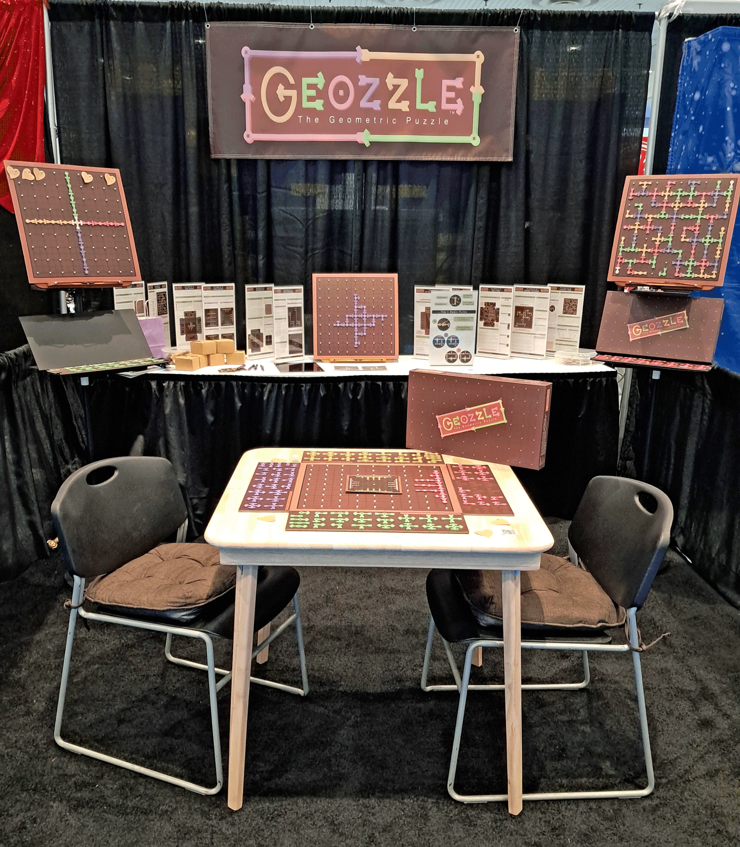 A booth display for Geozzle, a geometric puzzle game, with signs and game boards on tables and black curtains as background.