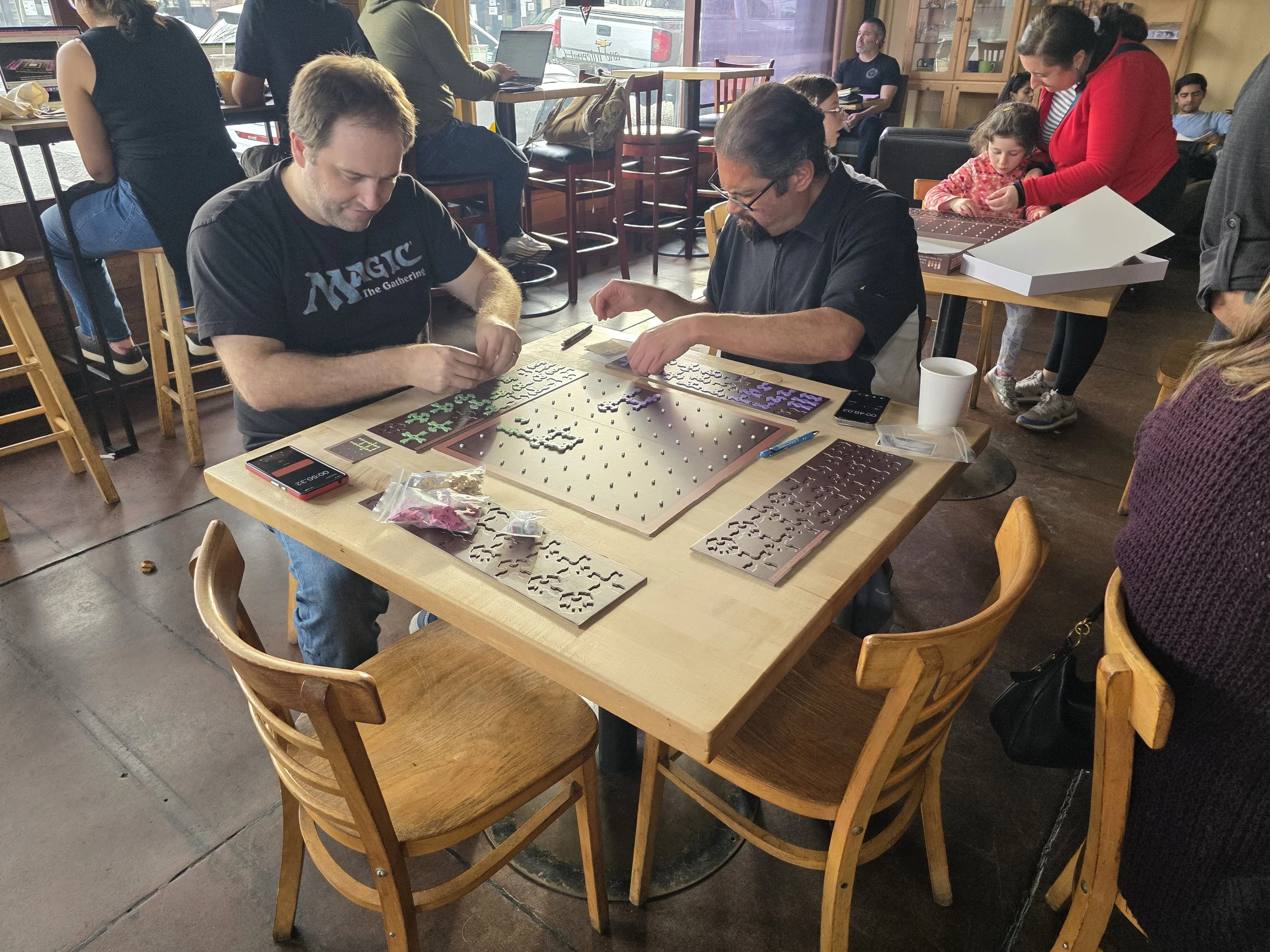 Two men are playing a puzzle game at a wooden table in a busy cafe, with some puzzle pieces on the table and a smartphone beside them. Multiple people are seated and working or socializing inside the cafe.