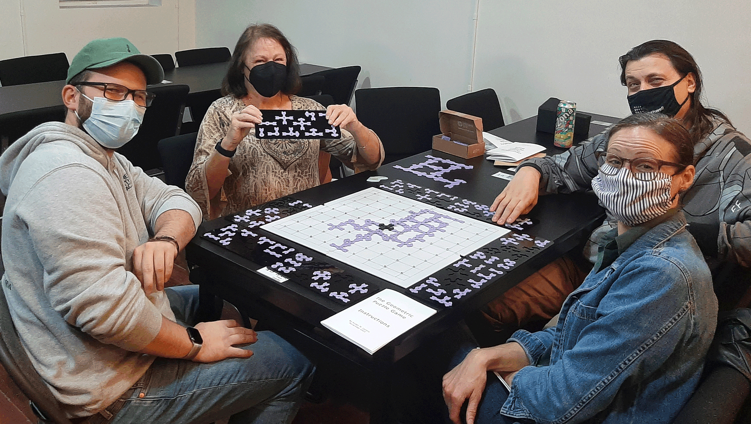 Group of five people sitting around a table playing a geometric puzzle game, with a game board and puzzle pieces. All individuals are wearing masks and casual clothing.