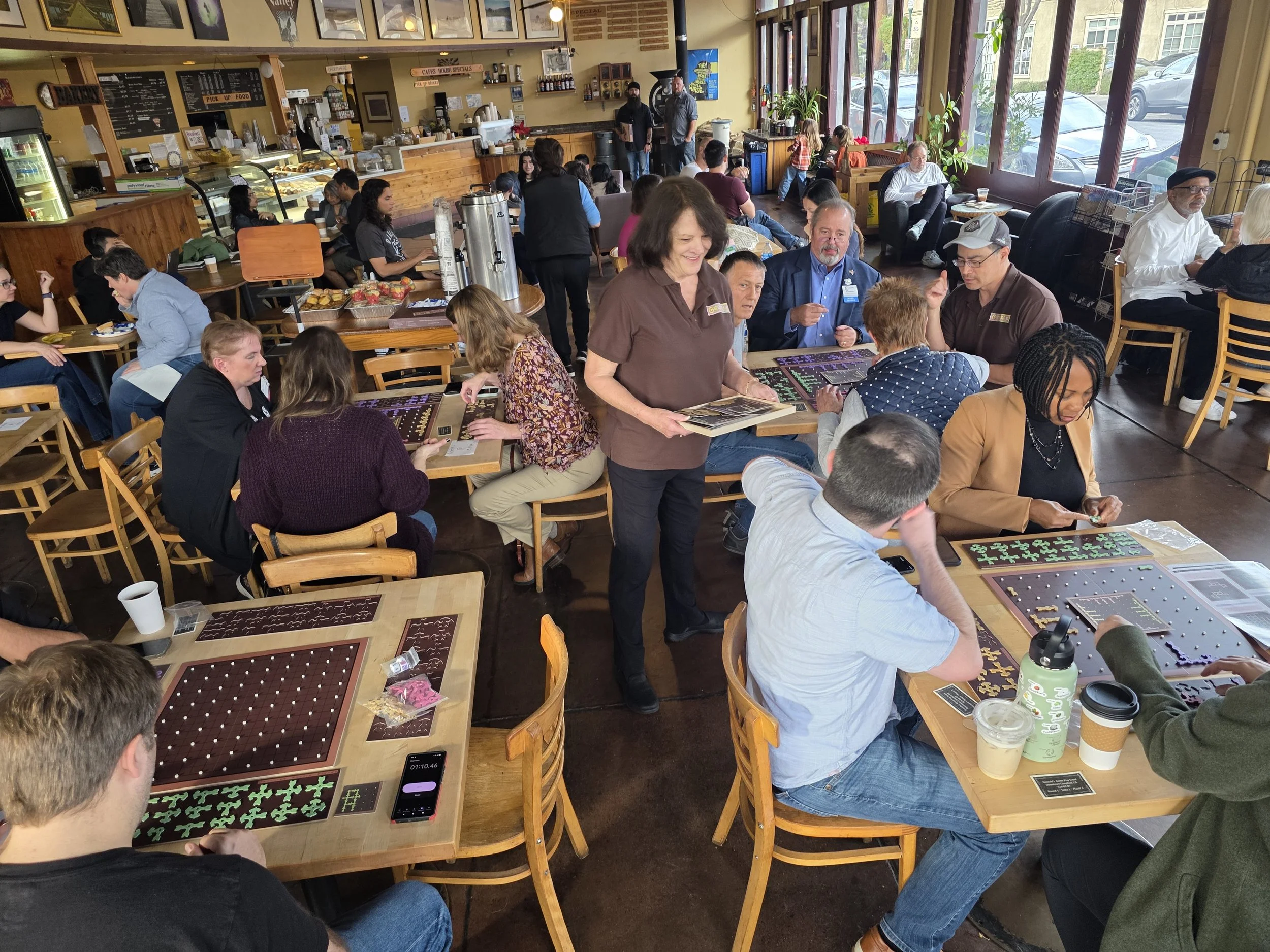 People playing board games at tables inside a busy cafe or coffee shop with large windows and a counter area.