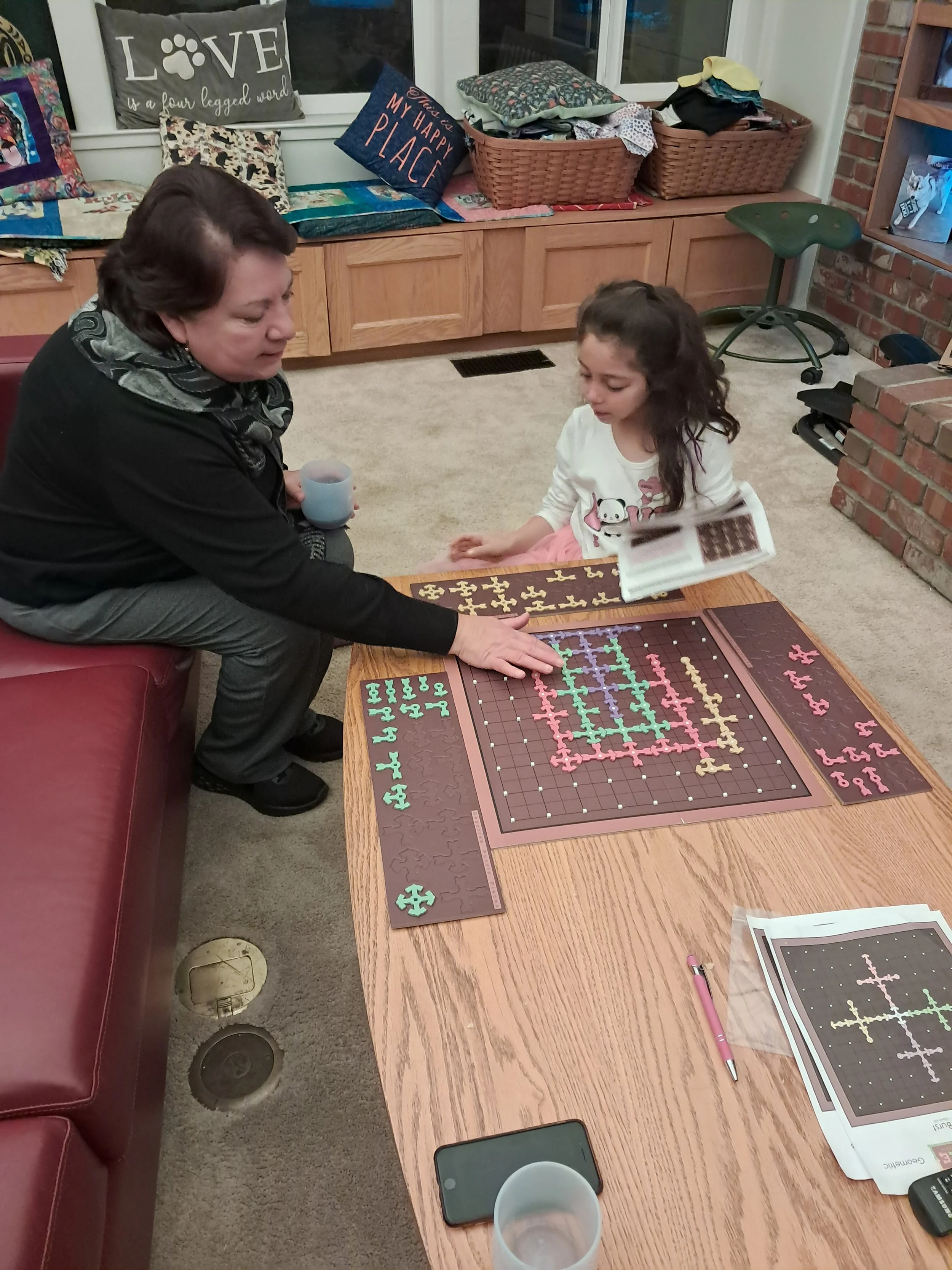 A woman and a girl are sitting at a wooden table playing a puzzle game with a puzzle board and pieces. The table has a cup, a smartphone, a pink pen, and a printed game sheet. In the background, there are pillows on a window seat, baskets with clothes, a green stool, and a brick fireplace.