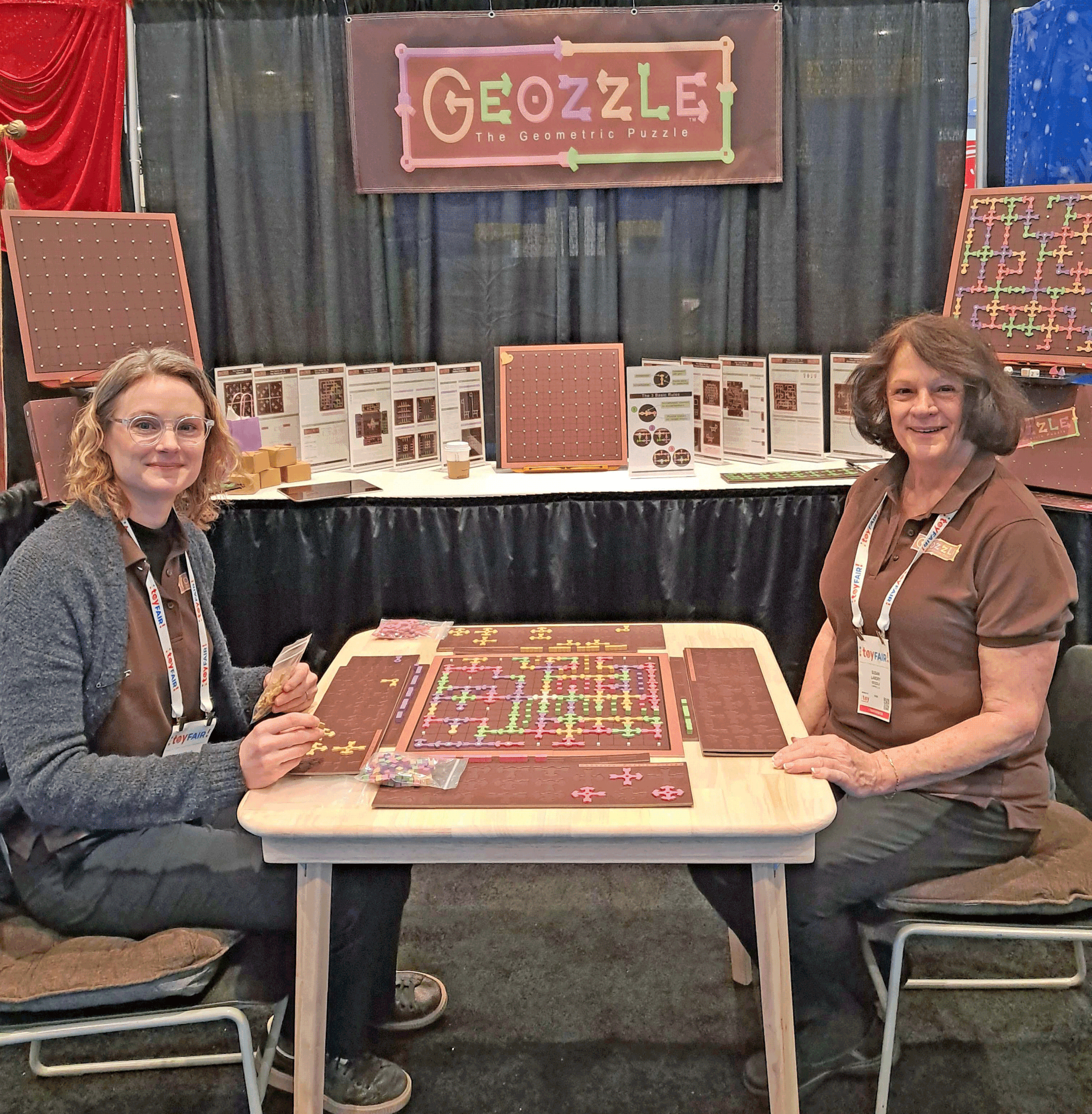 At Toy Fair, two women are seated at a table displaying a geometric puzzle called Geozzle, with various puzzle pieces and informational brochures in the background. The display features a sign reading 'GEOZZLE The Geometric Puzzle' and decorative puzzle pieces.
