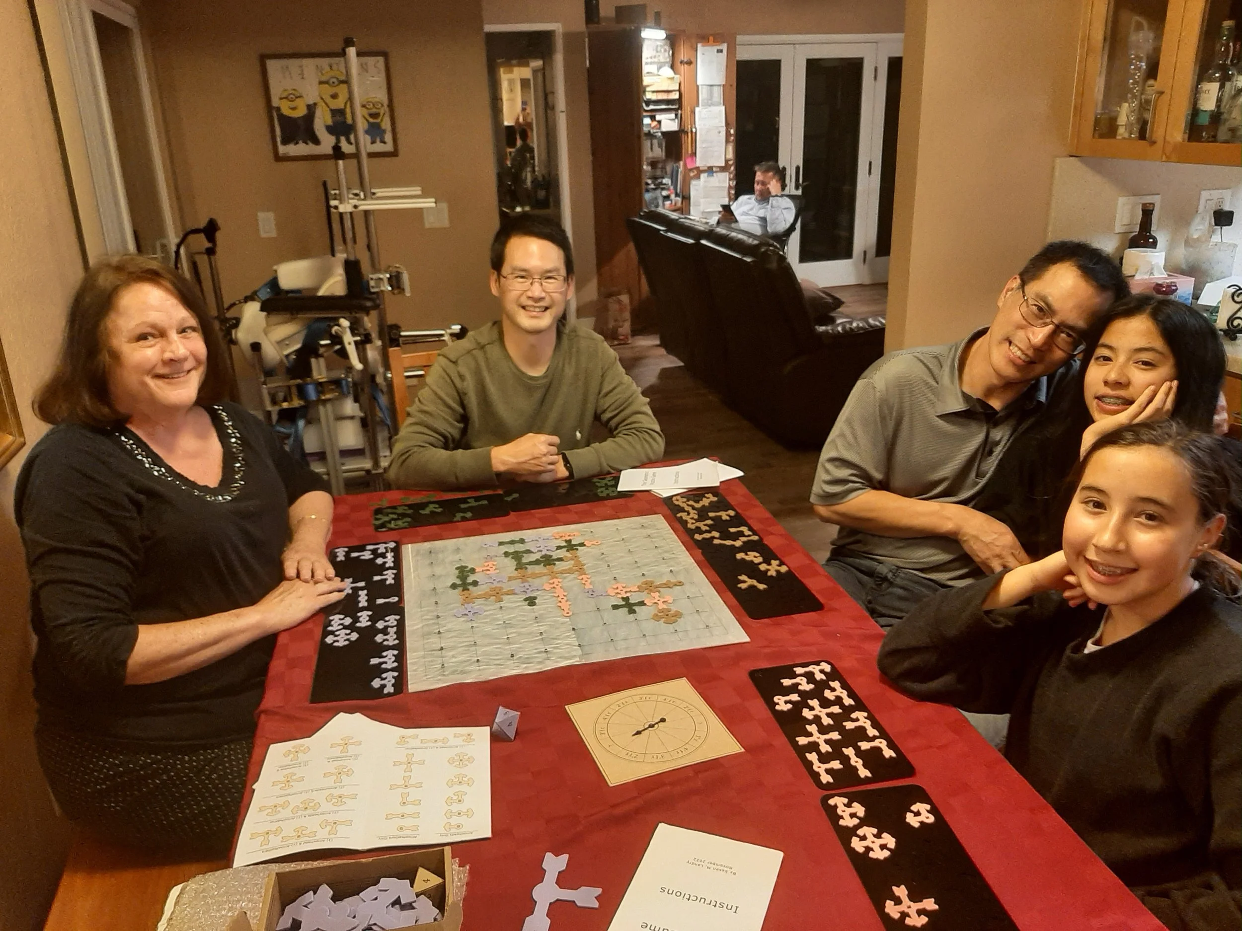A group of five people gathered around a table playing a GEOZZLE  puzzle, with completed puzzle pieces and game materials on the table, in a cozy home setting.