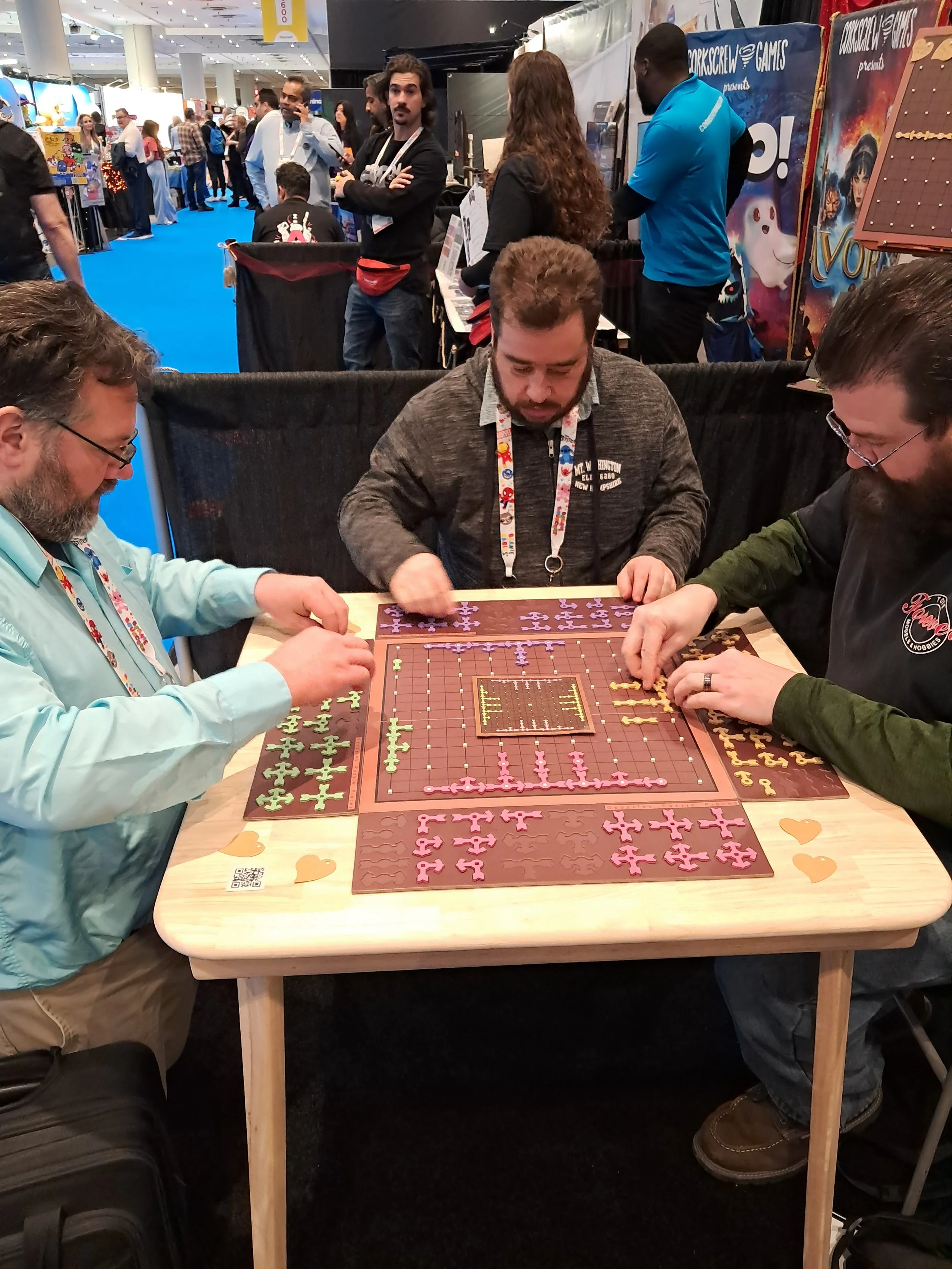 People playing a tabletop board game at a convention or event, with vendors and attendees visible in the background.