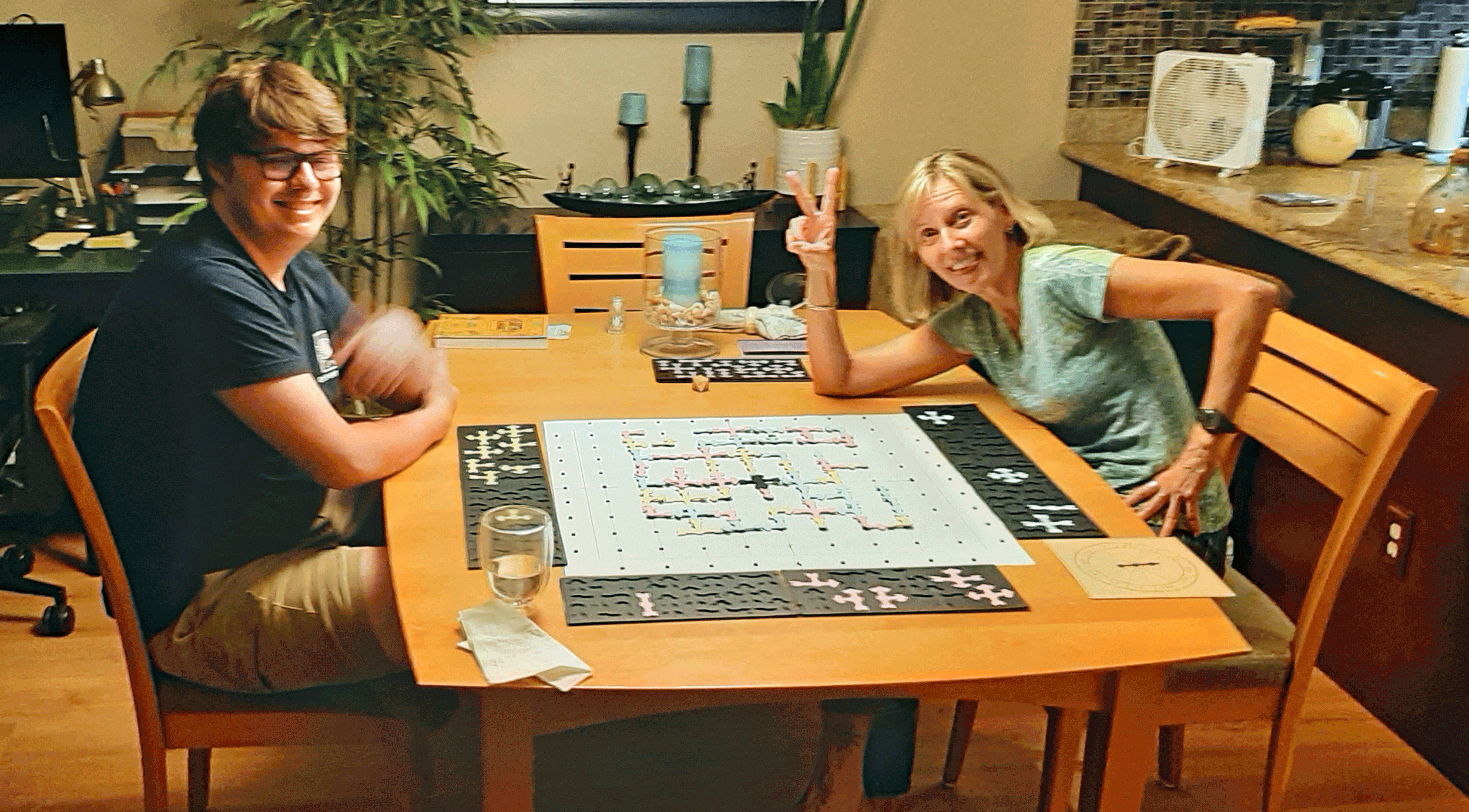 Two women sitting at a dining table playing a board game. One woman is making a peace sign with her fingers. The table has a game board with puzzle pieces, glasses of water, and various small items. In the background, there are plants, a fan, and kitchen items.